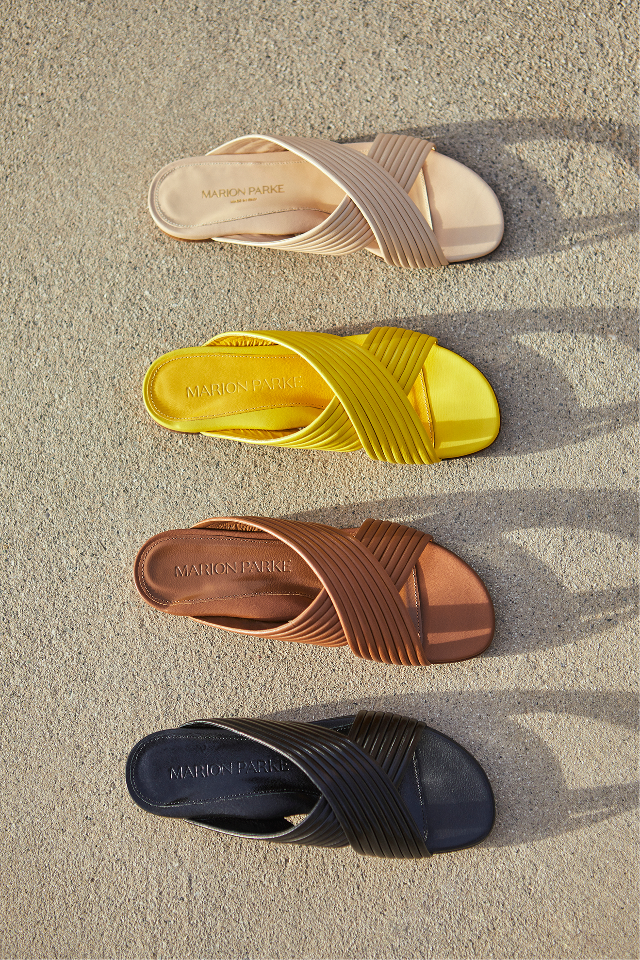 Four pairs of colorful sandals arranged on sand, each with crossed straps and the brand name 'Marion Parke' inside. The sandals are in beige, yellow, brown, and black.