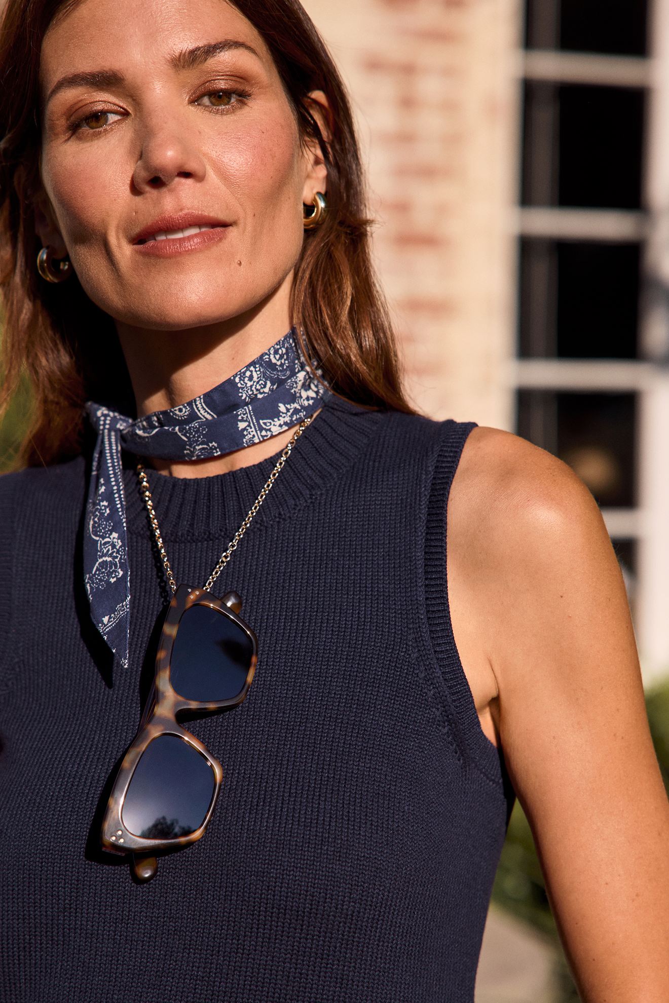 Close-up of a woman with brown hair, wearing gold hoop earrings, a blue patterned neck scarf, a navy sleeveless top, and tortoise shell sunglasses hanging from her shirt, standing outdoors with a blurred building in the background.