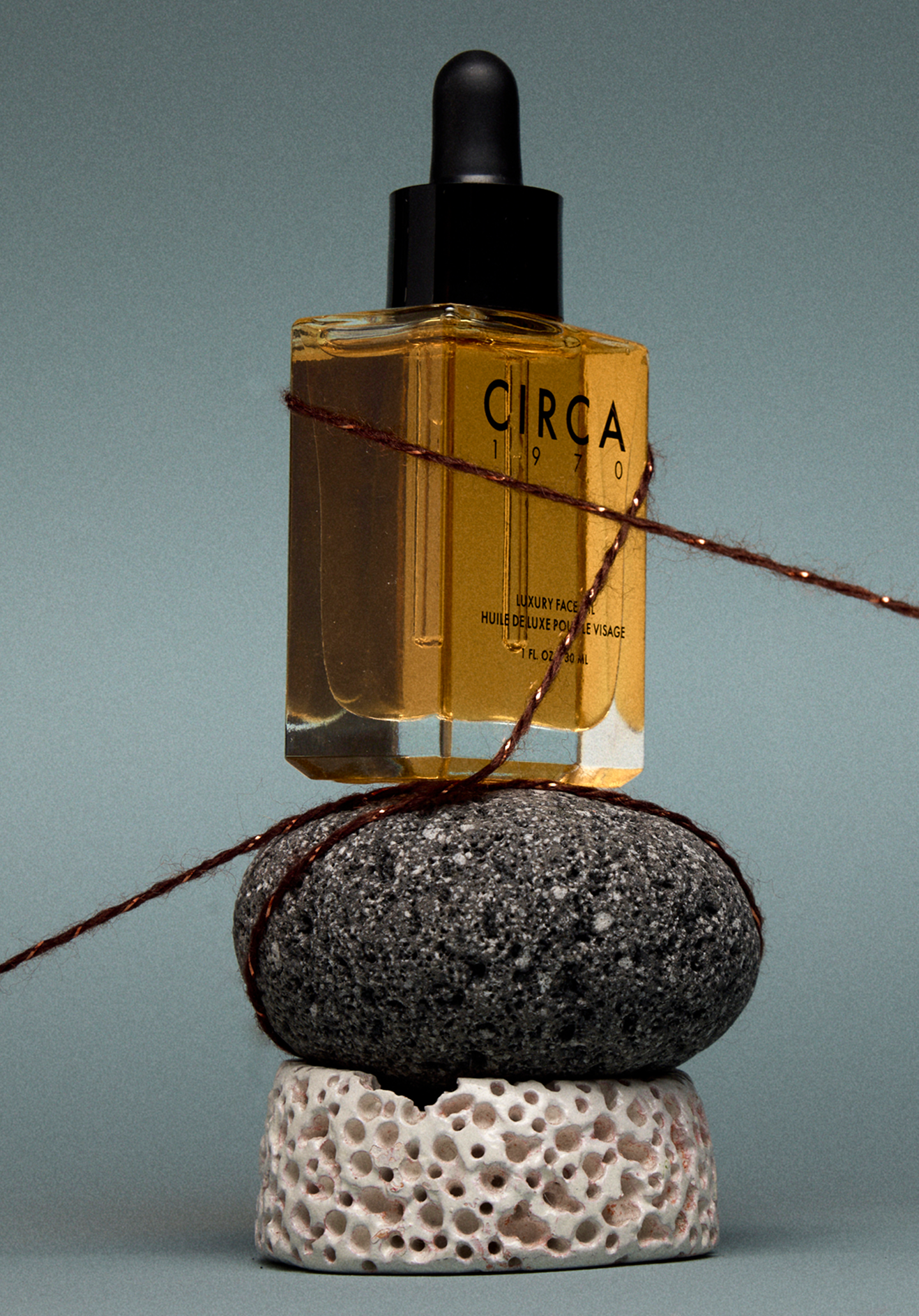 Stack of four different textured stones with a small, transparent, amber-colored bottle of face oil labeled 'CIRCA 1970' balanced on the top, connected by a piece of twine.