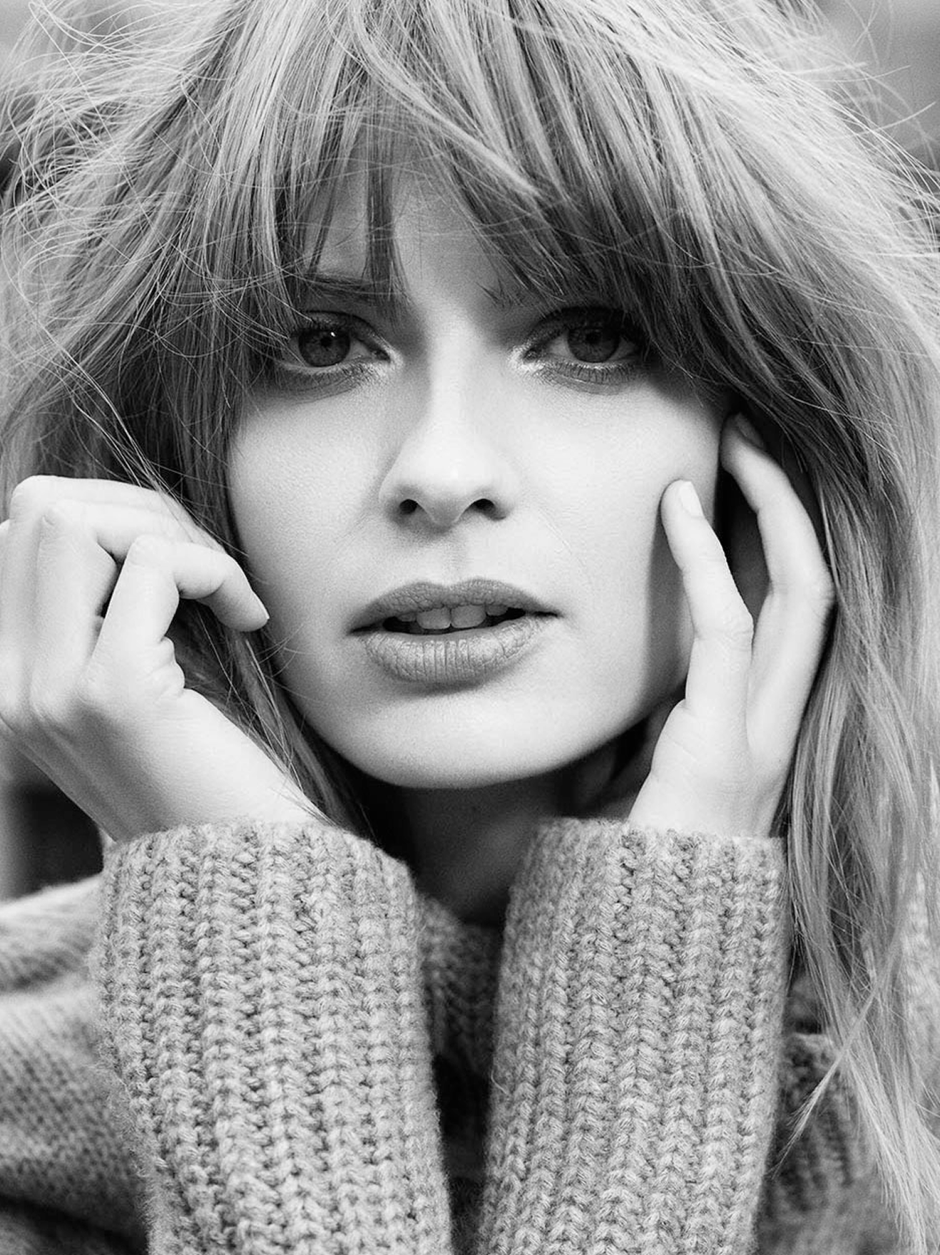 Black and white close-up portrait of a woman with wavy hair, touching her face with both hands, wearing a knitted sweater.