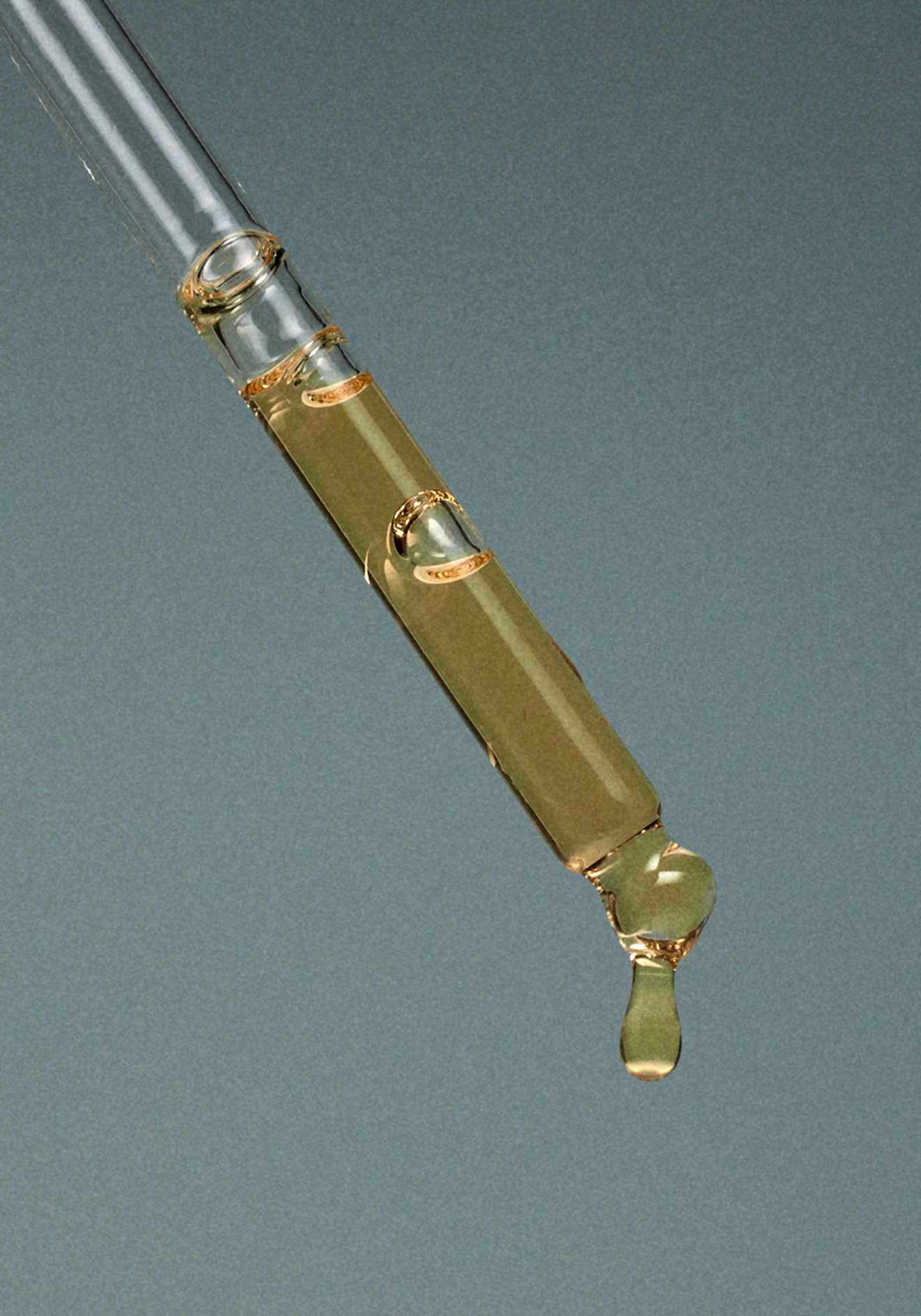 Close-up of a glass dropper with a small amount of clear liquid, with a droplet falling from the tip against a neutral background.