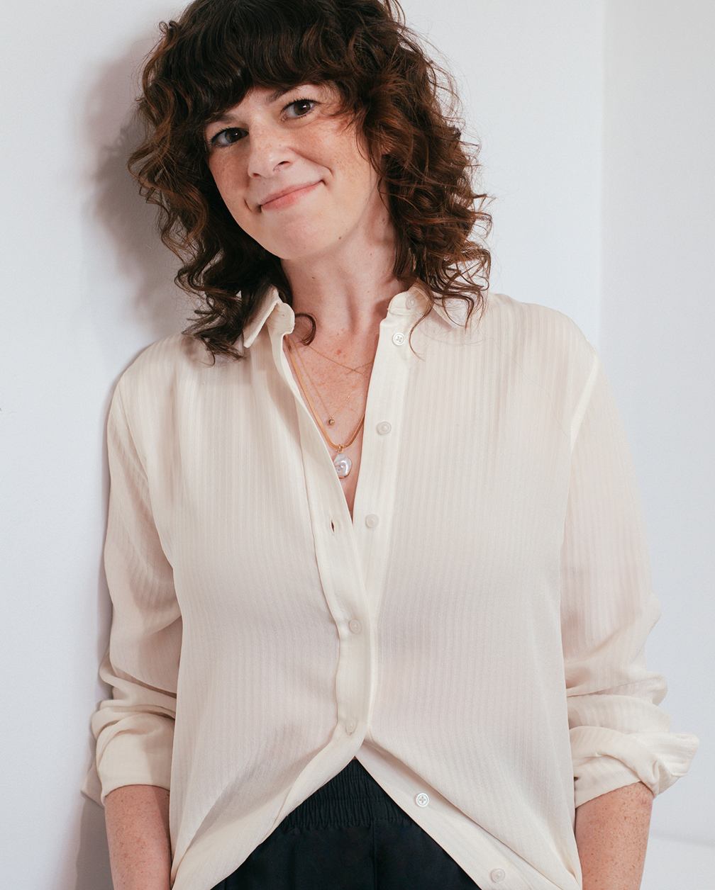 A woman with curly brown hair wearing a cream-colored blouse, layered necklaces, smiling softly and leaning against a white wall.