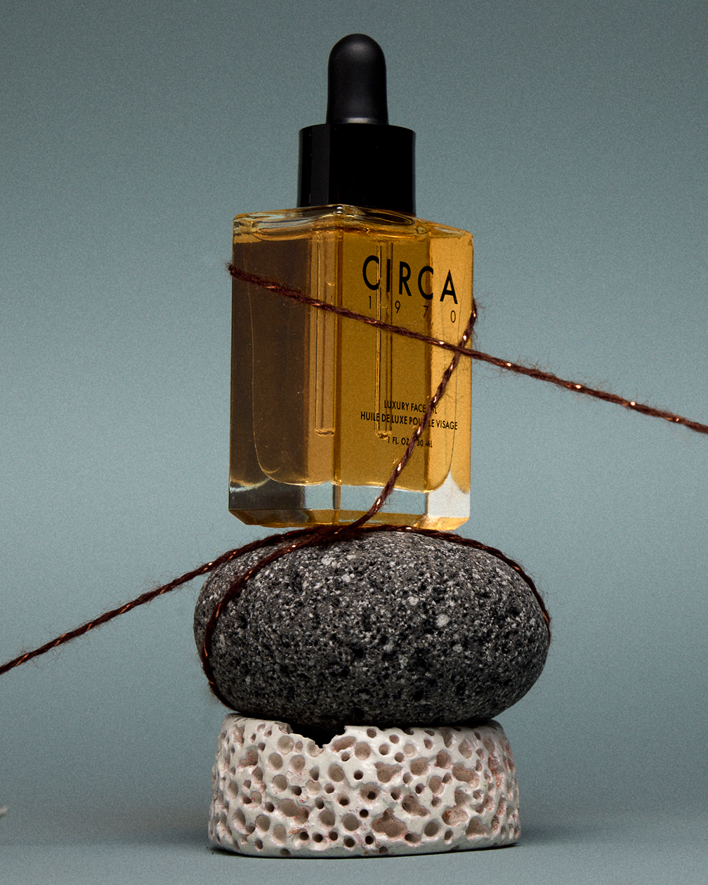 A glass bottle of Circa luxury face oil balanced on a stack of three rocks, with a string wrapped around the bottle and rocks, against a plain background.