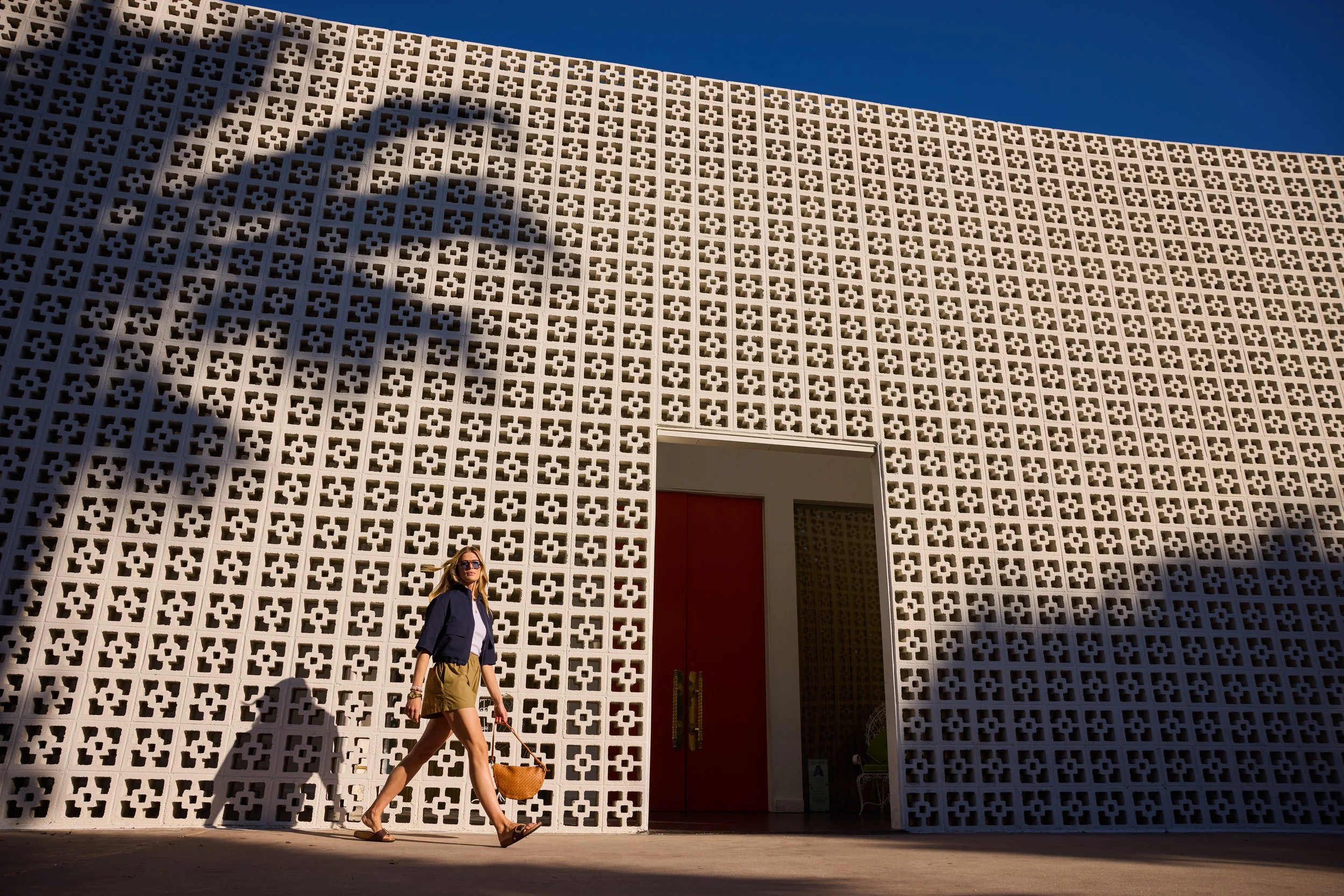 A woman walking in front of a beige geometric patterned building wall under a clear blue sky, carrying a large woven bag.