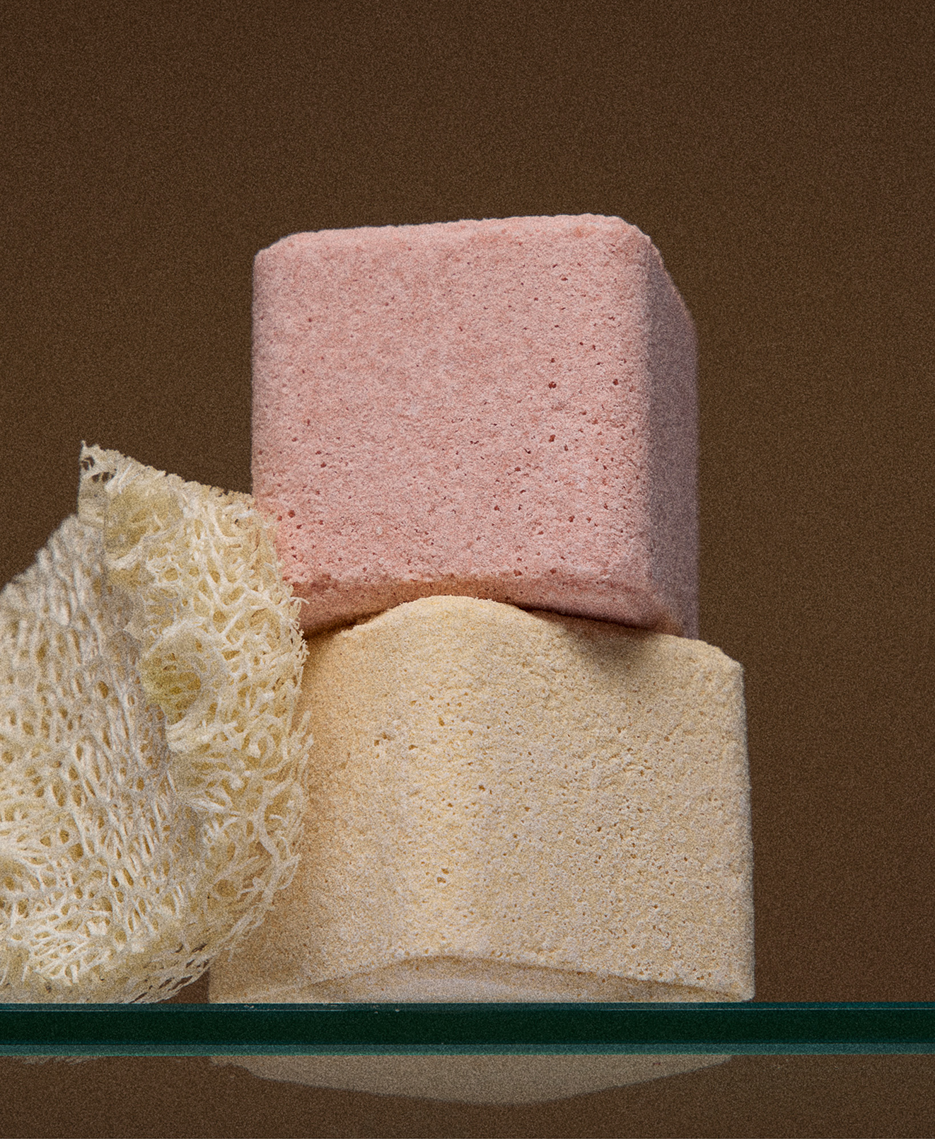 Three stacked spa or bath products in pink, yellow, and off-white colors with a white loofah sponge on the left side against a brown background.