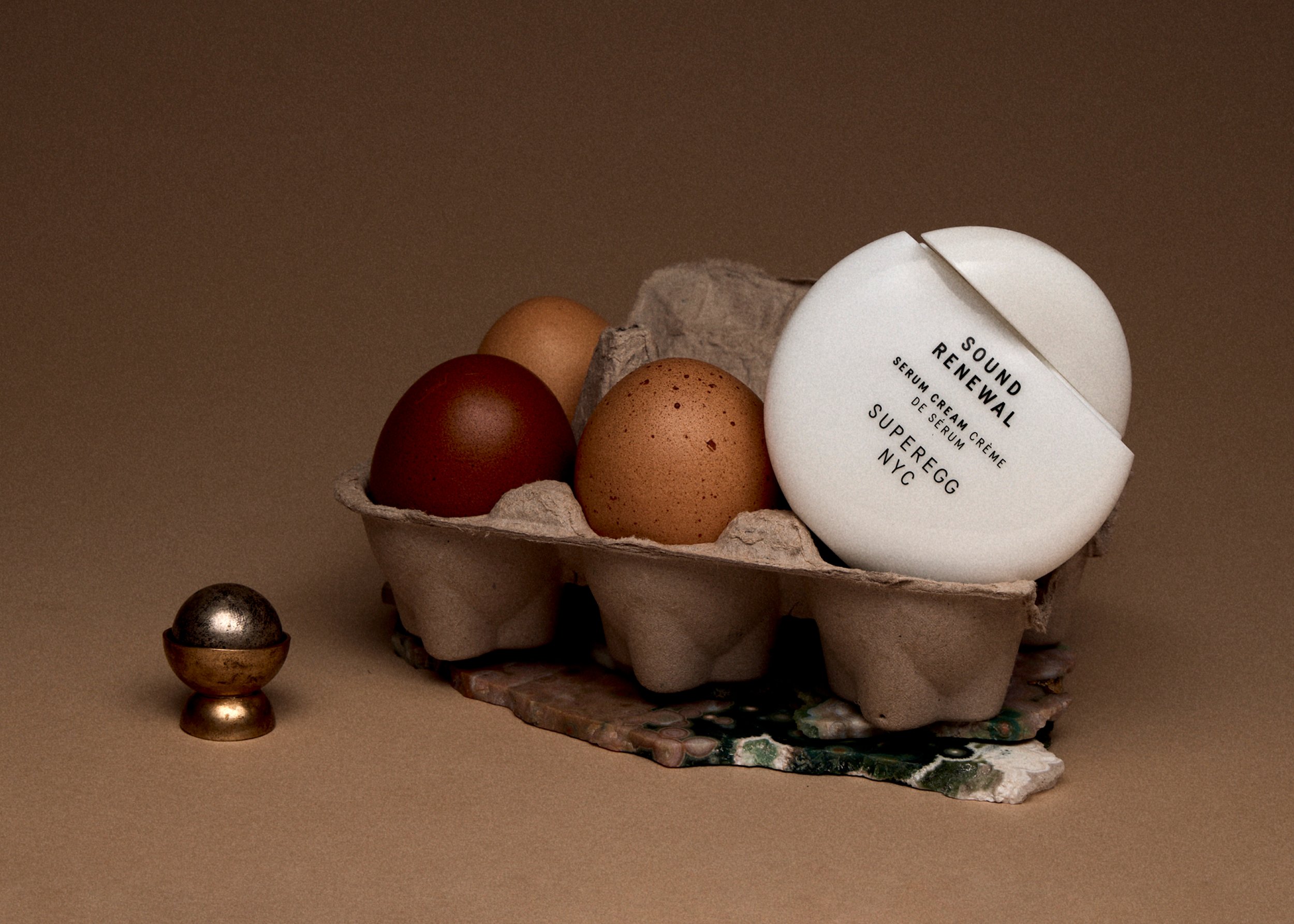 Eggs in a carton with a white container labeled 'Sound Renewal Serum' placed among them, with a few more small metallic decorative objects nearby, set against a neutral background.