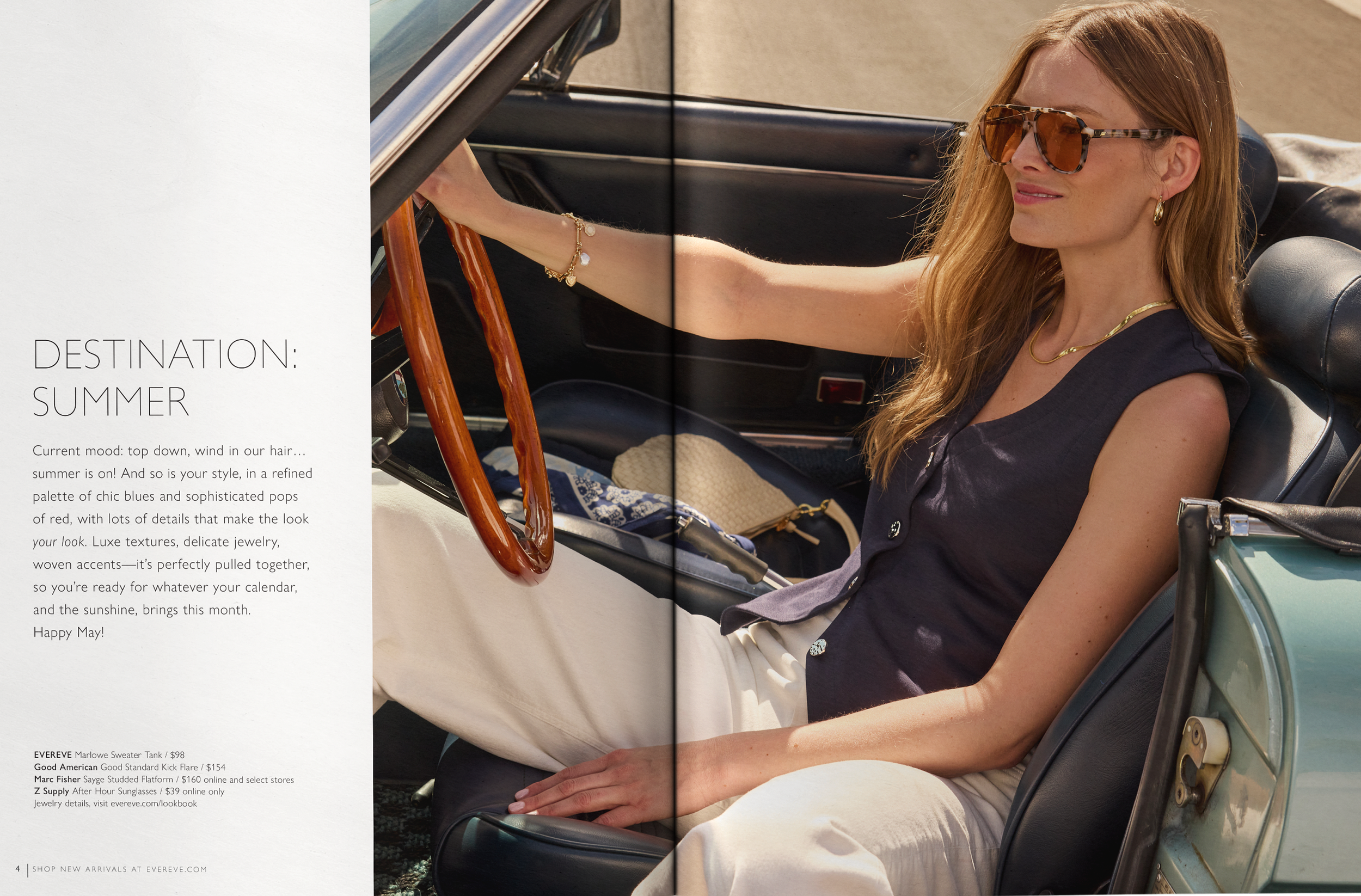 A woman with red hair wearing sunglasses, gold jewelry, a navy sleeveless top, and white pants, sitting in a vintage convertible car with a wooden steering wheel and black interior. She is smiling and enjoying a sunny day.
