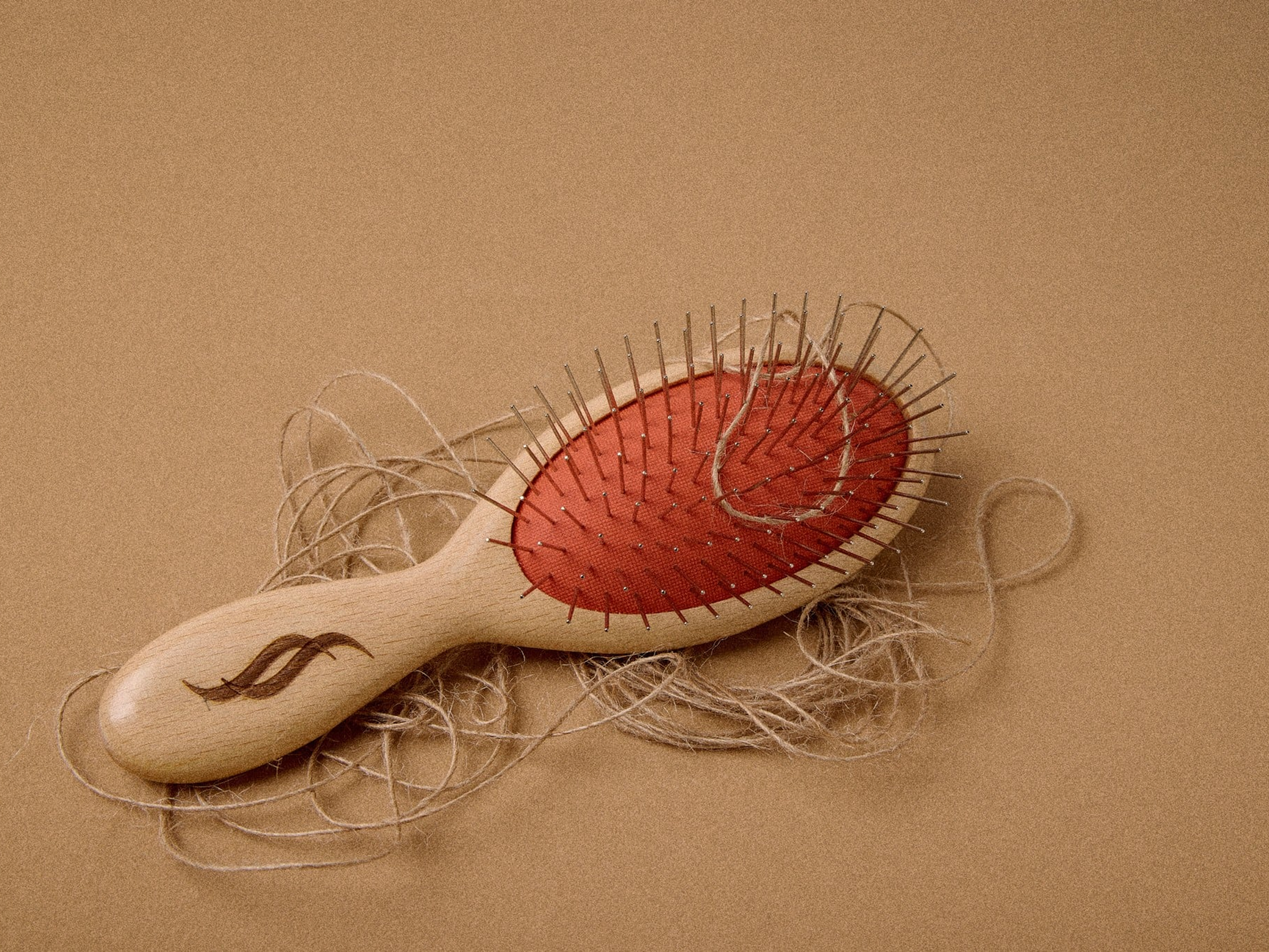 A hairbrush with a red rubber base and metal pins, placed on a beige background, with loose light-colored hair strands around it.