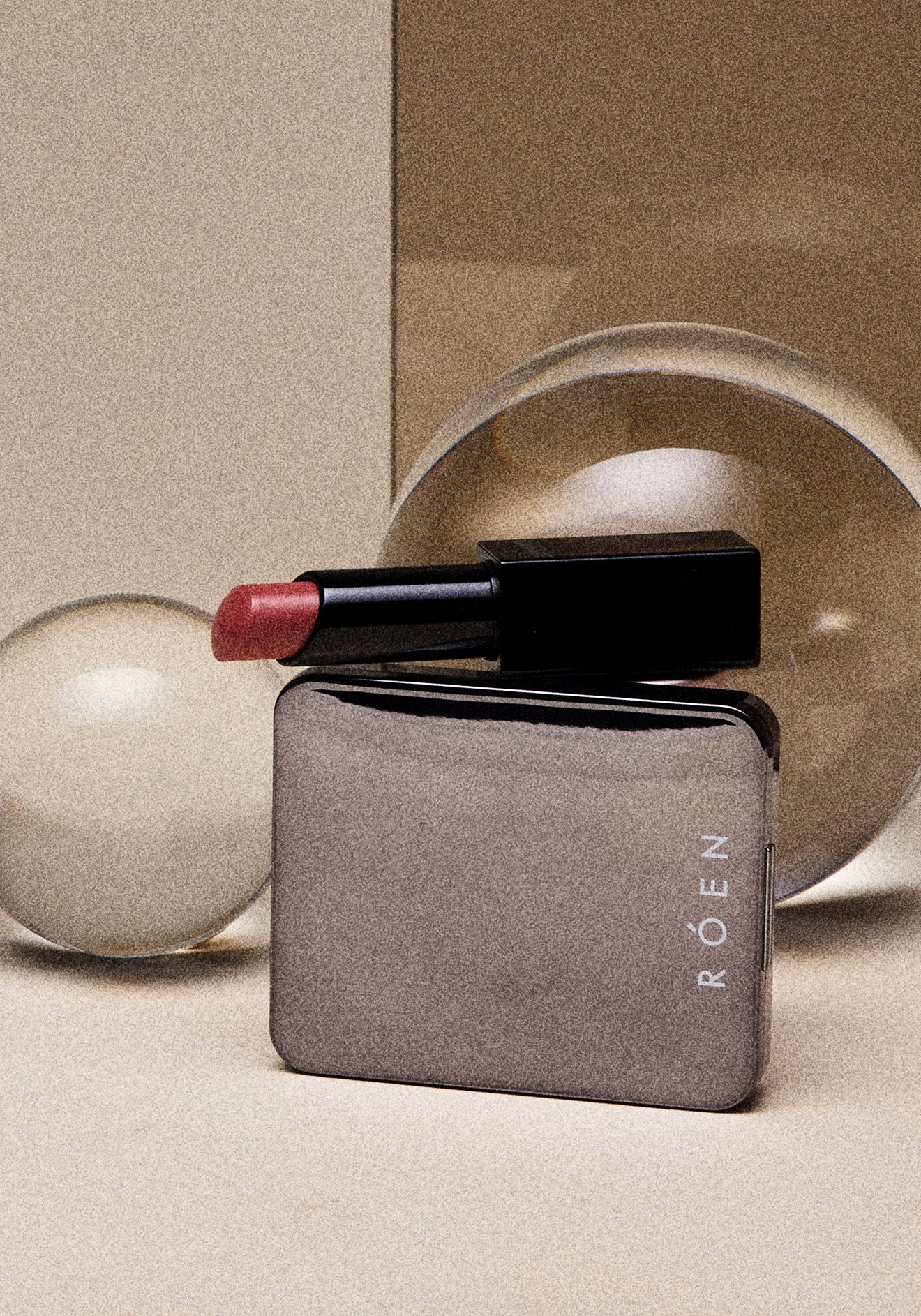 A lipstick with a muted red color on top of a gray compact case with the brand name 'ROËZEN' on it, against a beige background featuring round beige objects.