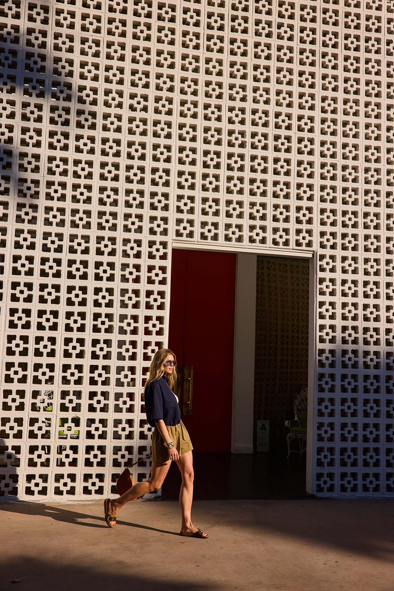 A woman wearing sunglasses, a navy blue jacket, and khaki shorts, walking on a sidewalk in front of a building with a decorative concrete block wall and a red door.
