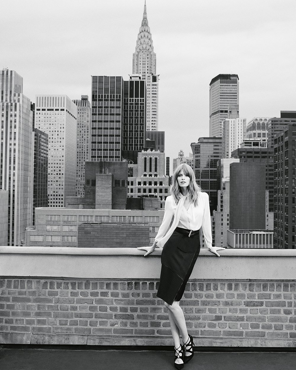 A woman in a white blouse and dark skirt standing on a rooftop with a city skyline, including the Empire State Building, in the background.