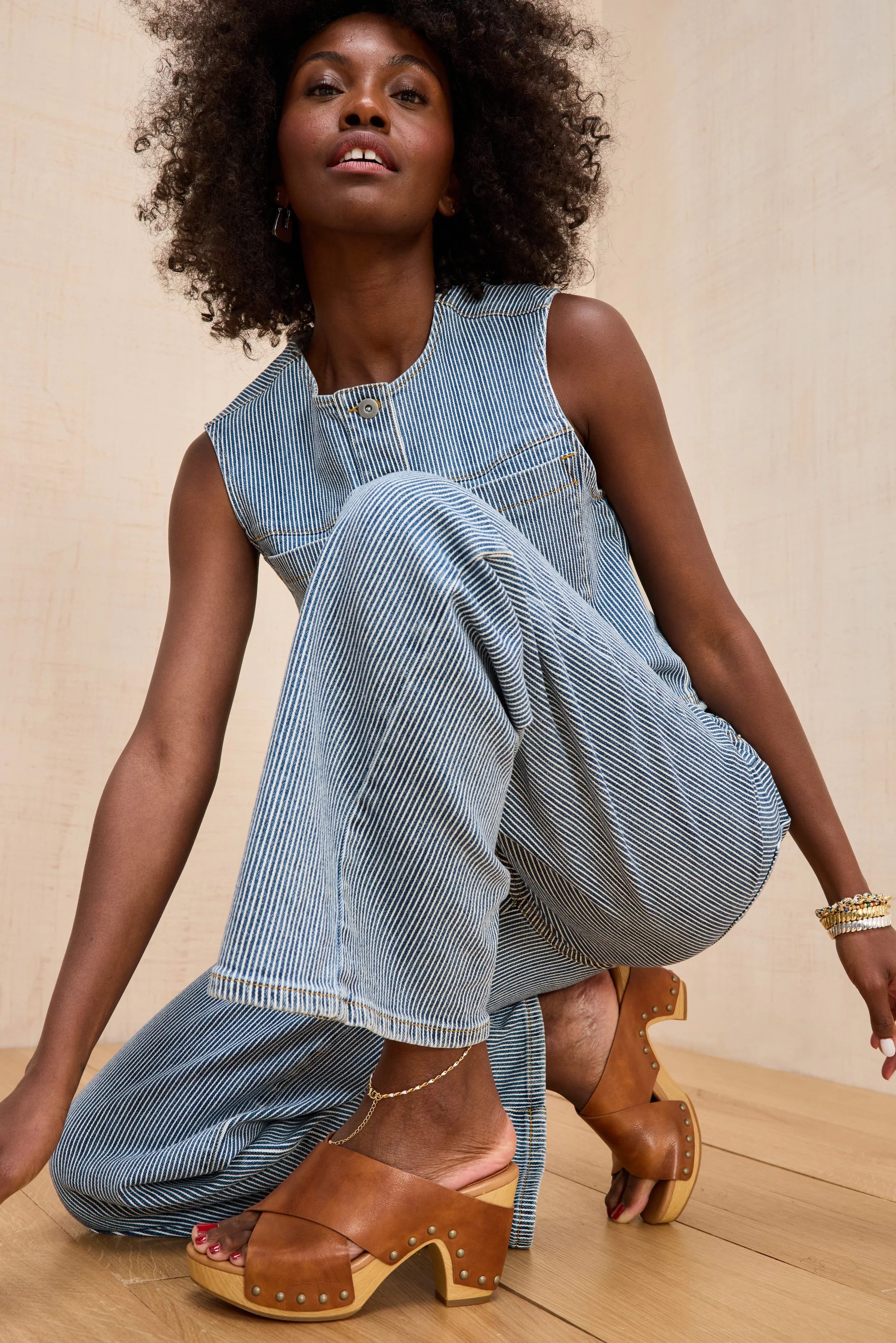 A woman with dark skin, curly hair, wearing a sleeveless striped jumpsuit, sitting on the floor and leaning back. She is wearing brown platform sandals and jewelry, with a plain light-colored background.