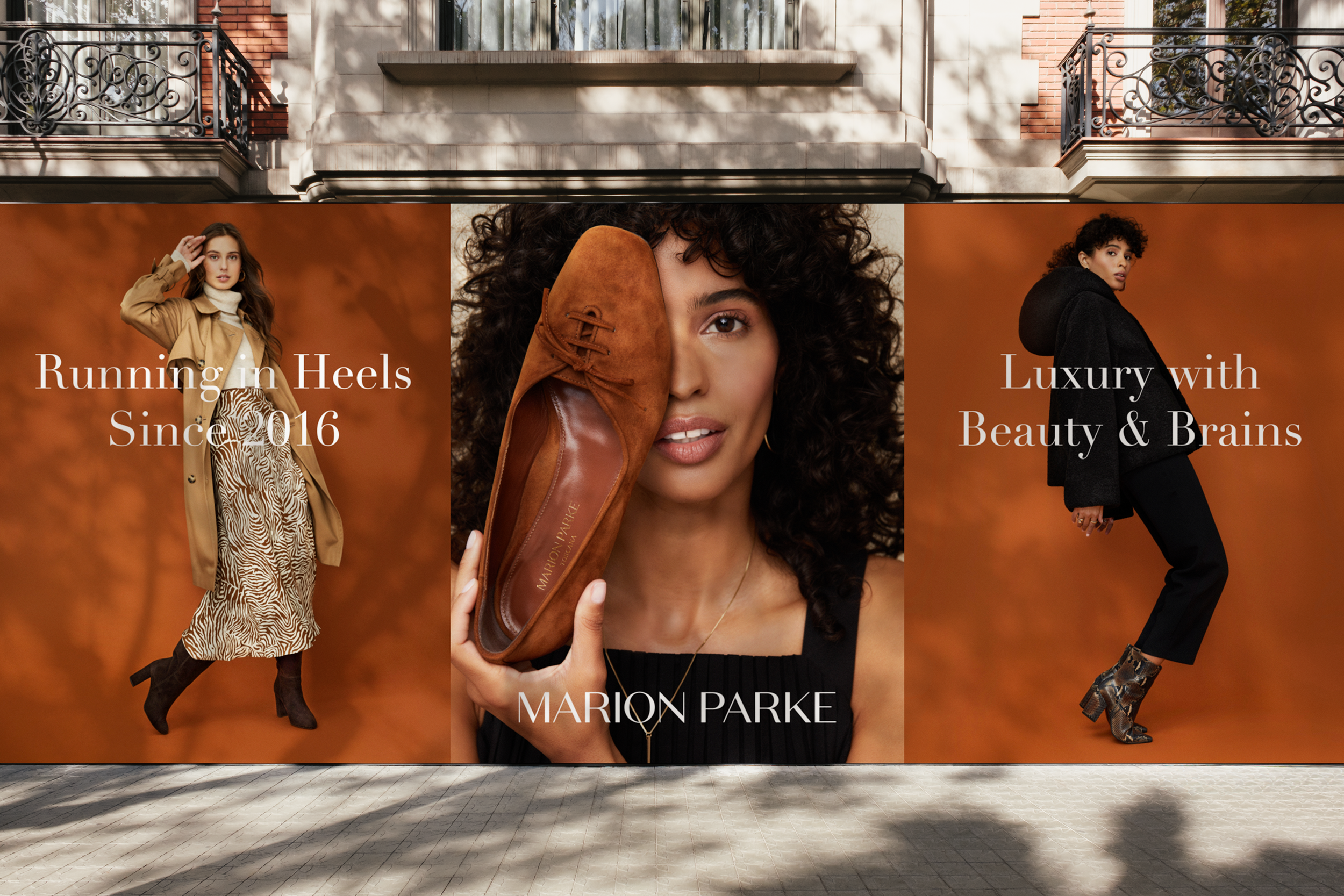 A large outdoor billboard with three images of women modeling fashion. The left woman is in a camel coat and patterned dress, the center woman has curly hair holding a brown shoe to her face, and the right woman wears a black hoodie and pants. The billboard features text promoting Marion Parke, a luxury shoes brand, with the phrases "Running in Heels Since 2016" and "Luxury with Beauty & Brains."