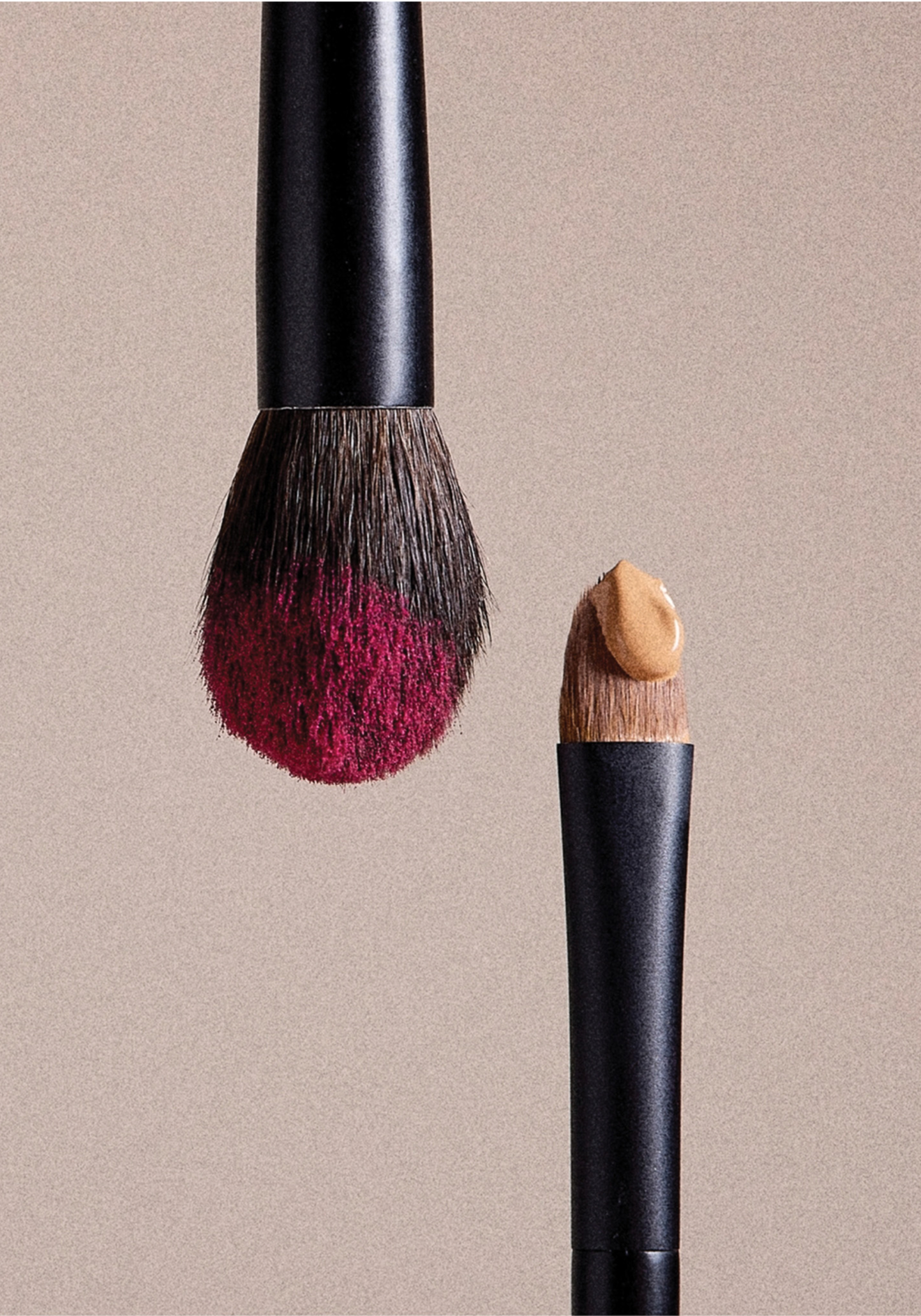 Close-up of a makeup brush with pink blush on the bristles and a small makeup sponge on the other brush handle, against a beige background.
