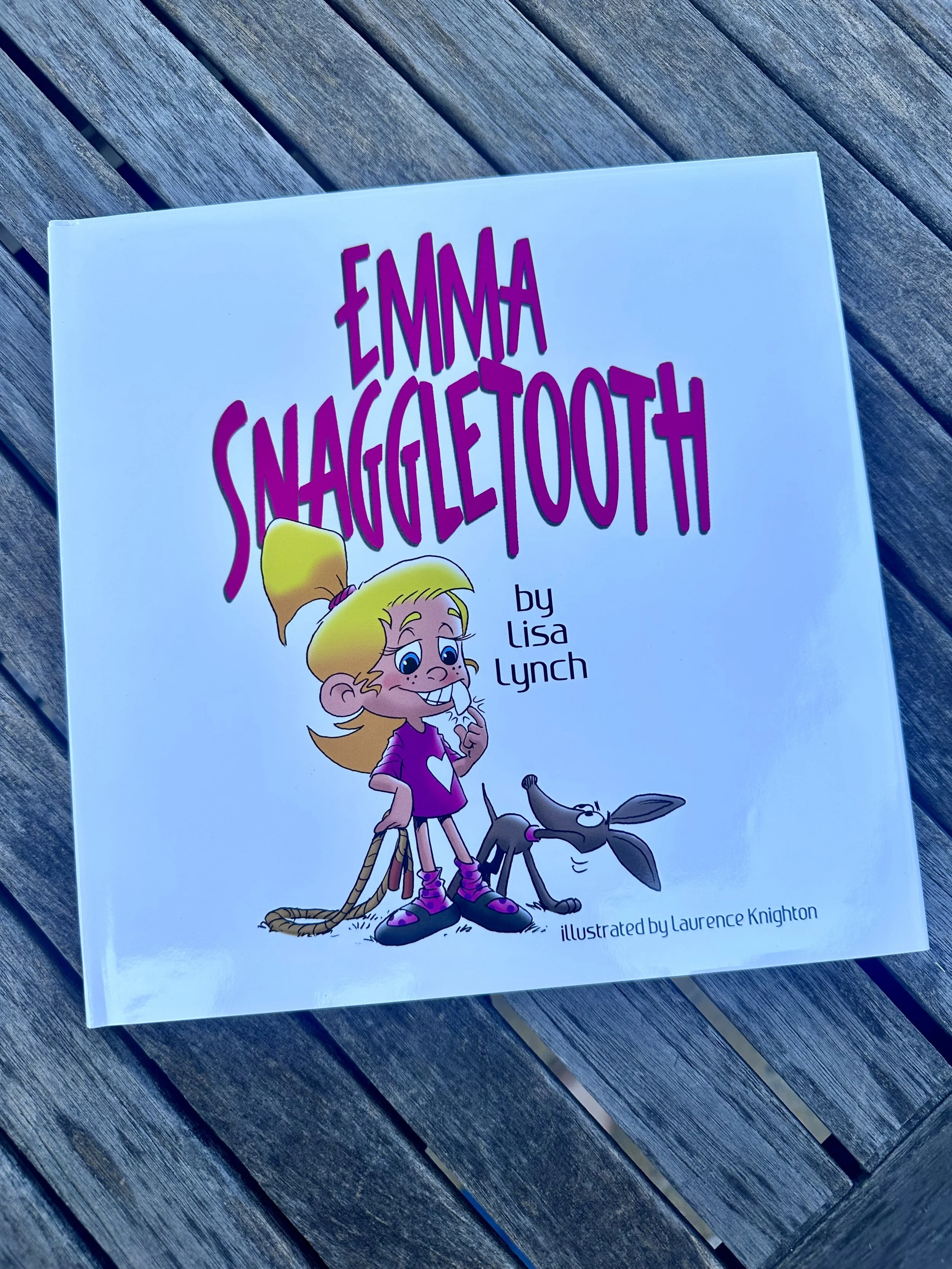 Book cover of "Emma Snaggletooth" by Lisa Lynch, illustrated by Laurence Knighton, featuring a cartoon girl with blonde hair, a pink shirt with a heart, and a dog on a wooden surface.