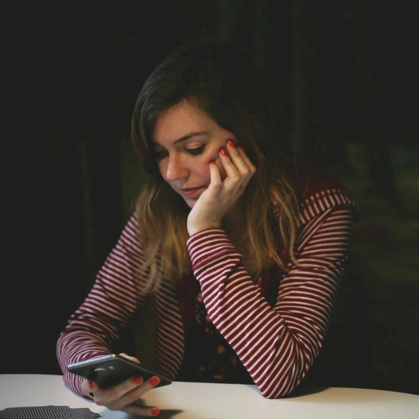 A young woman with light skin and long hair, wearing a red and white striped shirt, is sitting at a table and looking at her smartphone. She has red nail polish and appears to be deep in thought, resting her face on her hand.