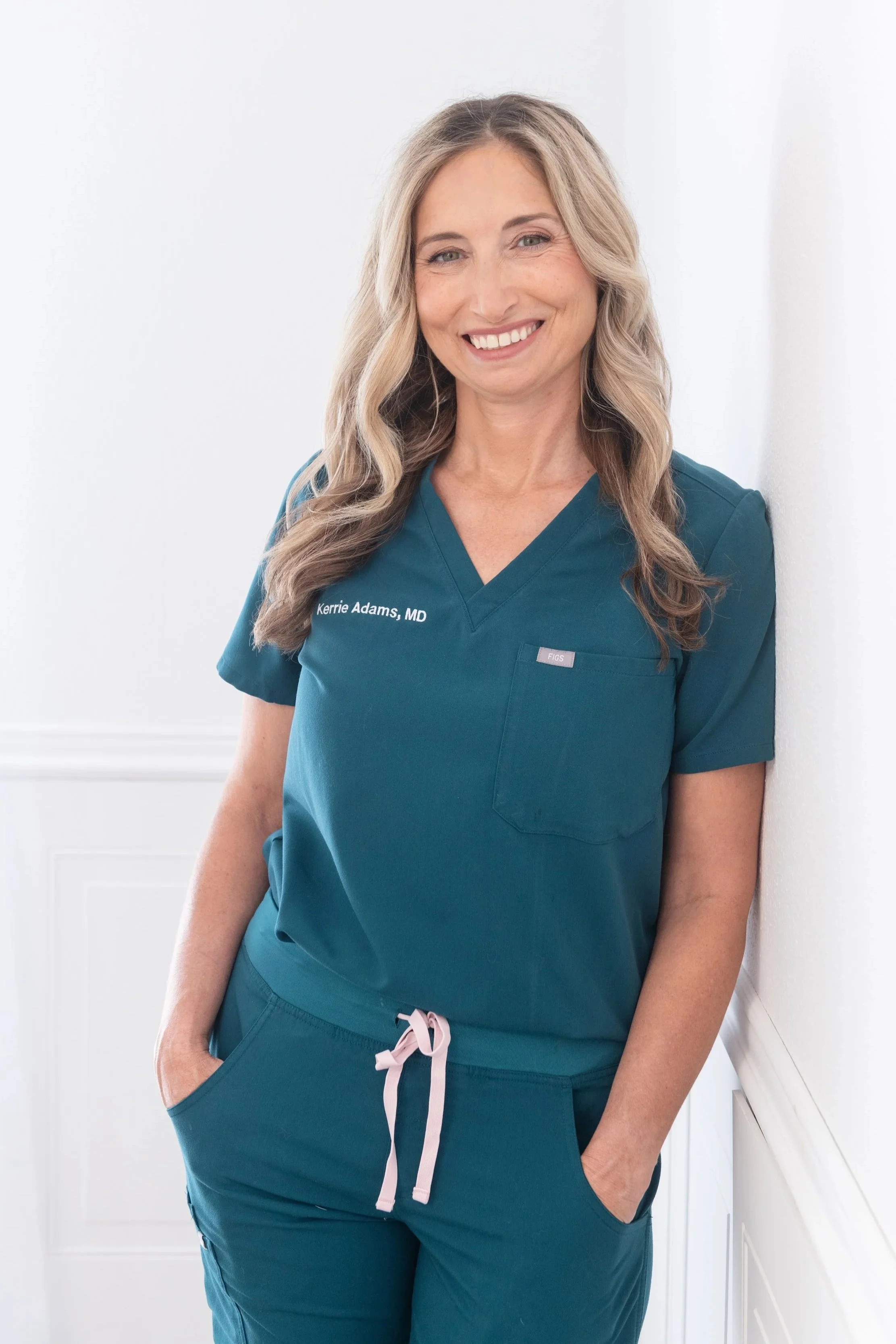 A woman wearing teal medical scrubs, smiling and leaning against a white wall.