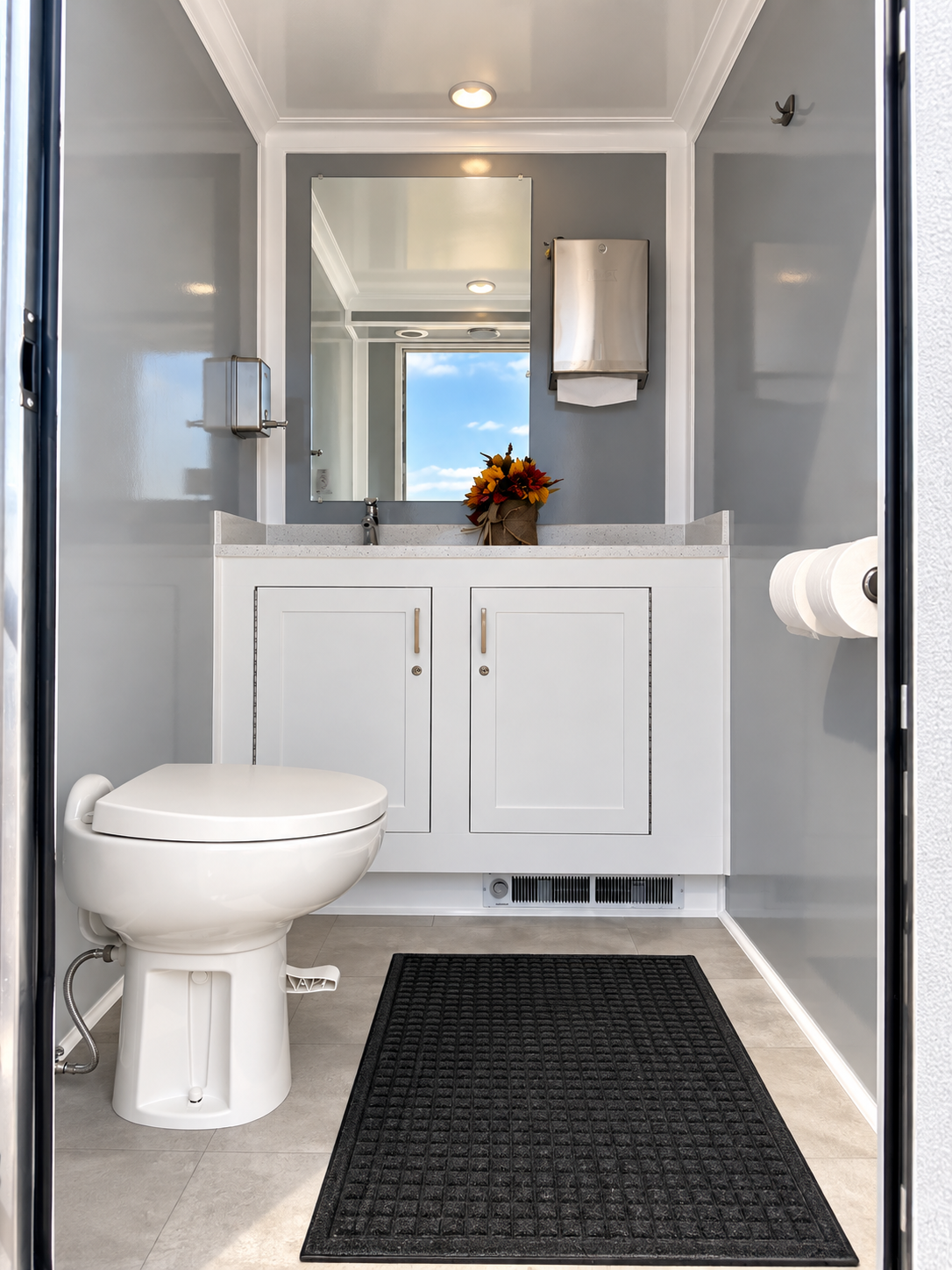 A small bathroom with a white toilet, a white vanity with a single sink, a mirror, and a small cabinet on the wall. There is a black rug in front of the vanity, a paper towel roll on a holder, and a decorative bouquet of flowers on the sink. The walls are painted light gray, and a window reflects the sky and clouds.