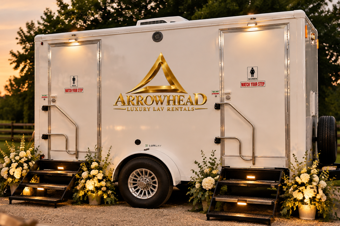 A white luxury lav rental trailer decorated with white flowers and green foliage, featuring the Arrowhead Luxury Lav Rentals logo in gold on its side, set outdoors at sunset.