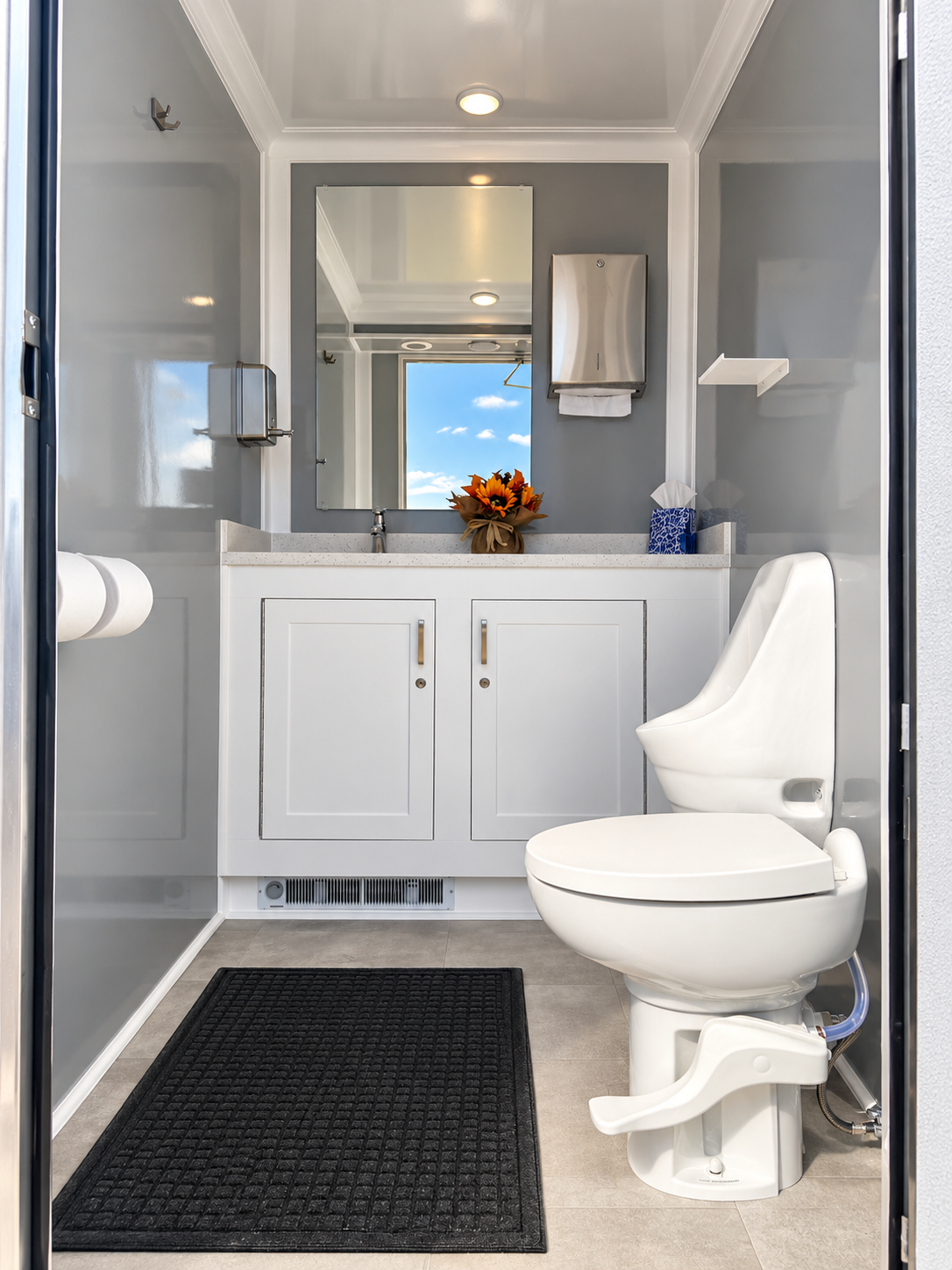 Small bathroom with a white toilet on the right, a white vanity with a mirror above it, a bouquet of flowers, a tissue box, and a soap dispenser. The room has gray walls, white trim, a black rug, and is well-lit with natural and ceiling lighting.