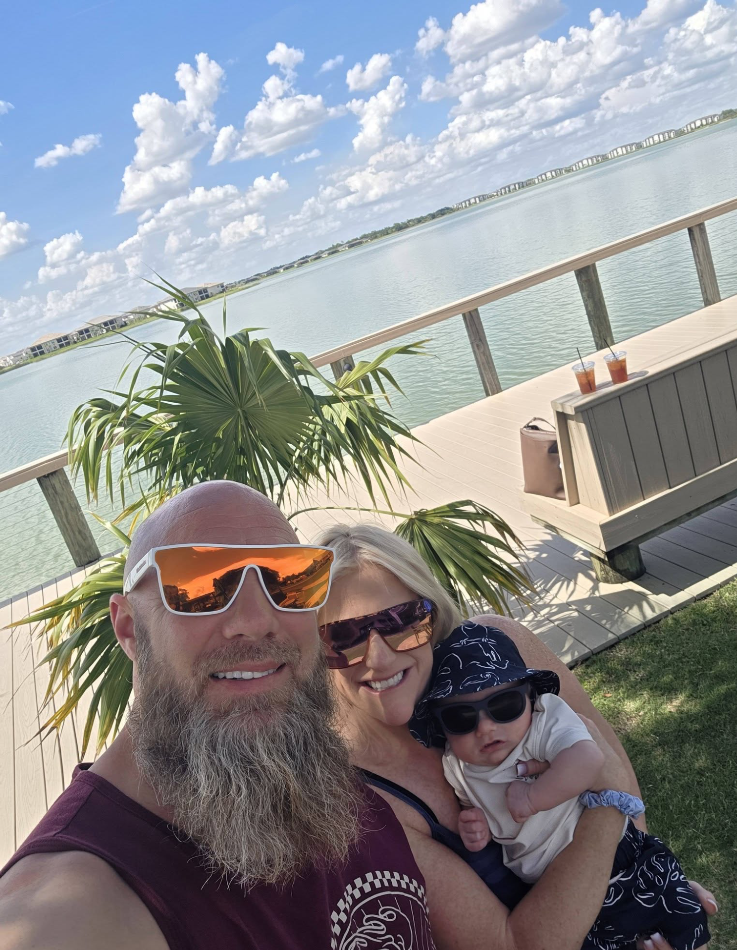 Smiling man with a beard and sunglasses, woman in sunglasses holding a child with sunglasses, all three enjoying sunny weather near water with a deck, railing, and clouds in the sky.