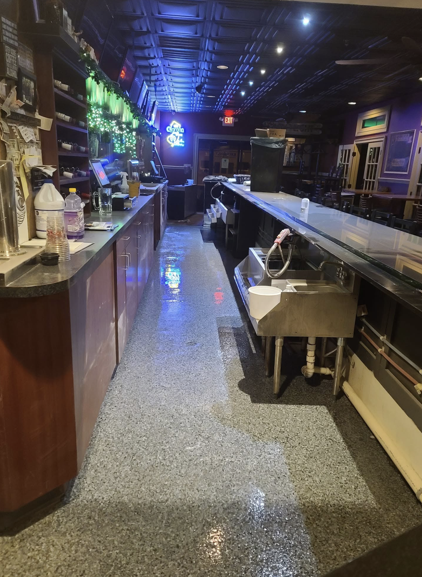Inside a bar, showing a long counter with drinks and supplies, a soda fountain, a sink, bar stools, and a dark ceiling with neon signs and decorative lighting.