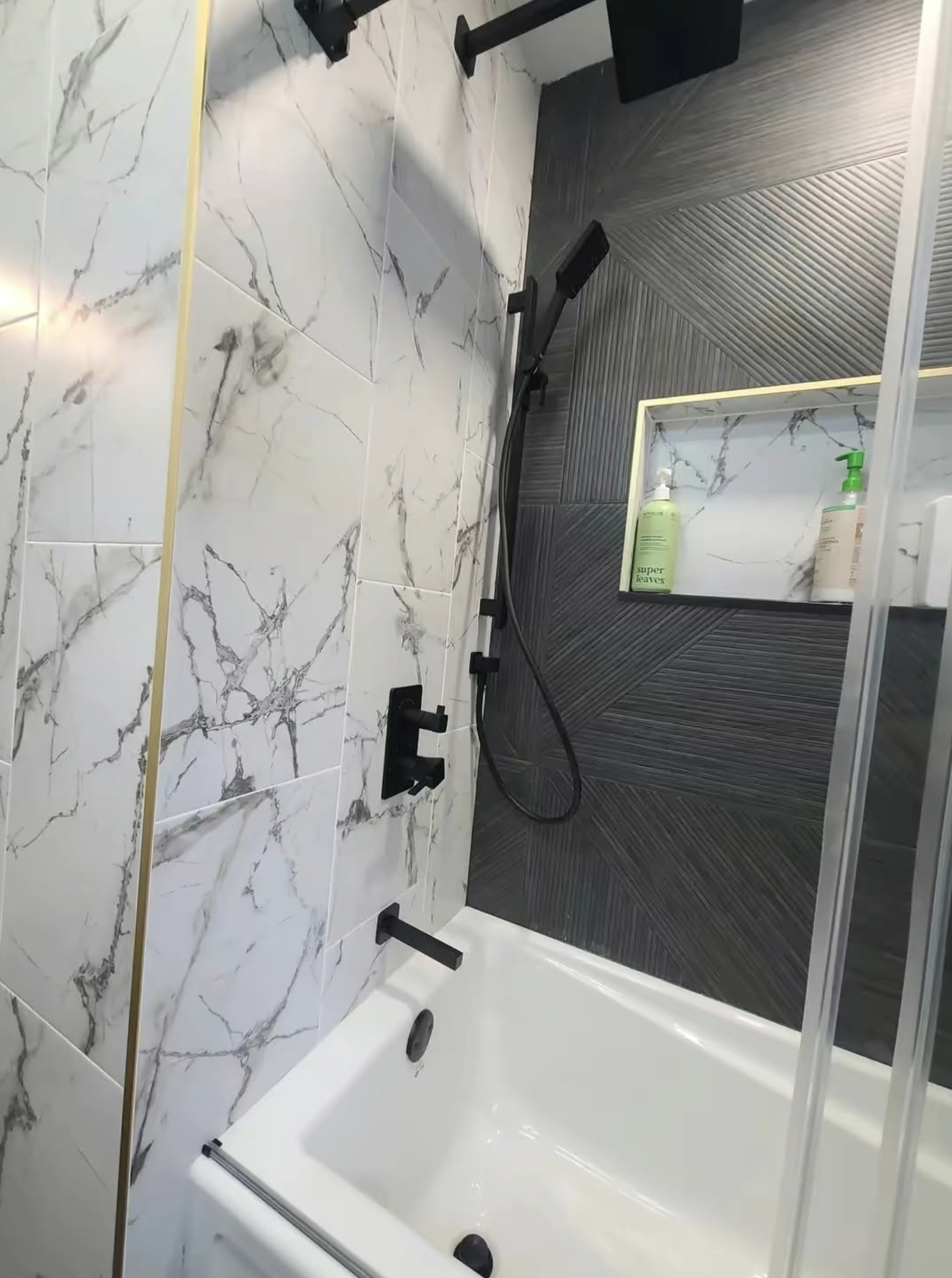 A modern bathroom shower with white marble tiles with gray veining on one wall and black textured tiles on the other wall. There are three black showerheads and a white shower base with a glass shower door.