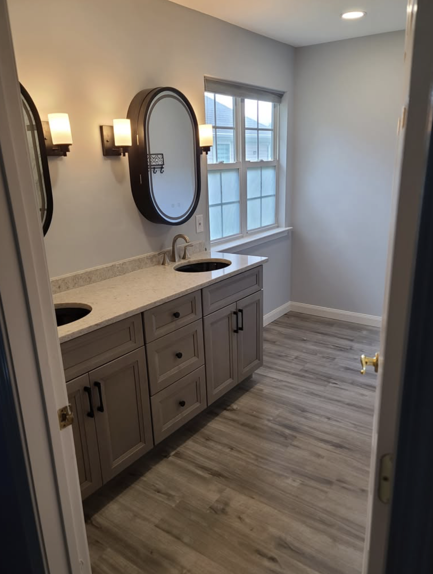 A bathroom with a double vanity, two black framed oval mirrors, wall-mounted lights, a window with blinds, light-colored cabinetry, and wood-look flooring.