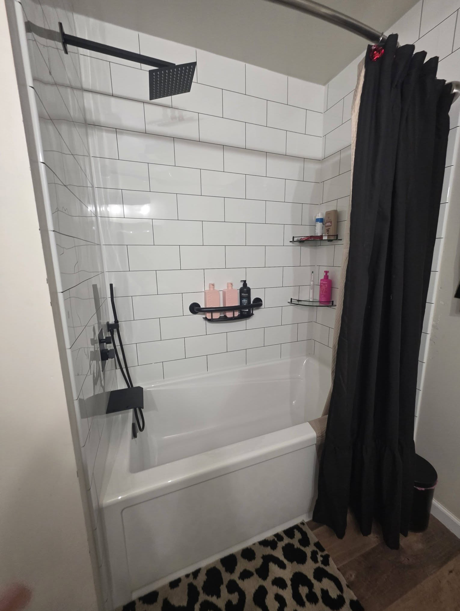 A bathroom shower with white subway tiles, a black shower curtain, and multiple shelves with personal care products.