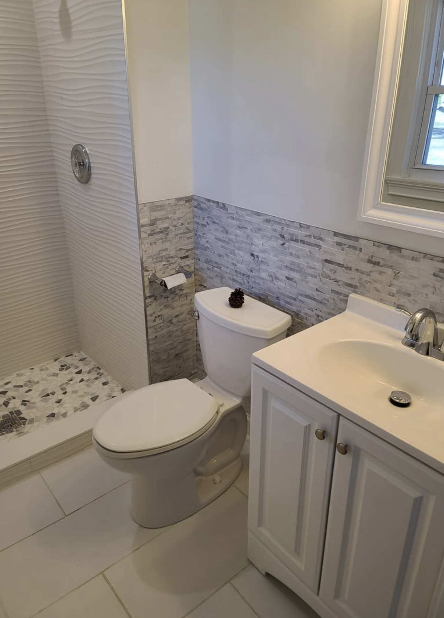Bathroom with a walk-in shower area, a toilet with a pine cone on top, and a sink with a white cabinet. The wall behind the toilet and sink has a decorative gray stone tile backsplash, and there is a window with white framing.