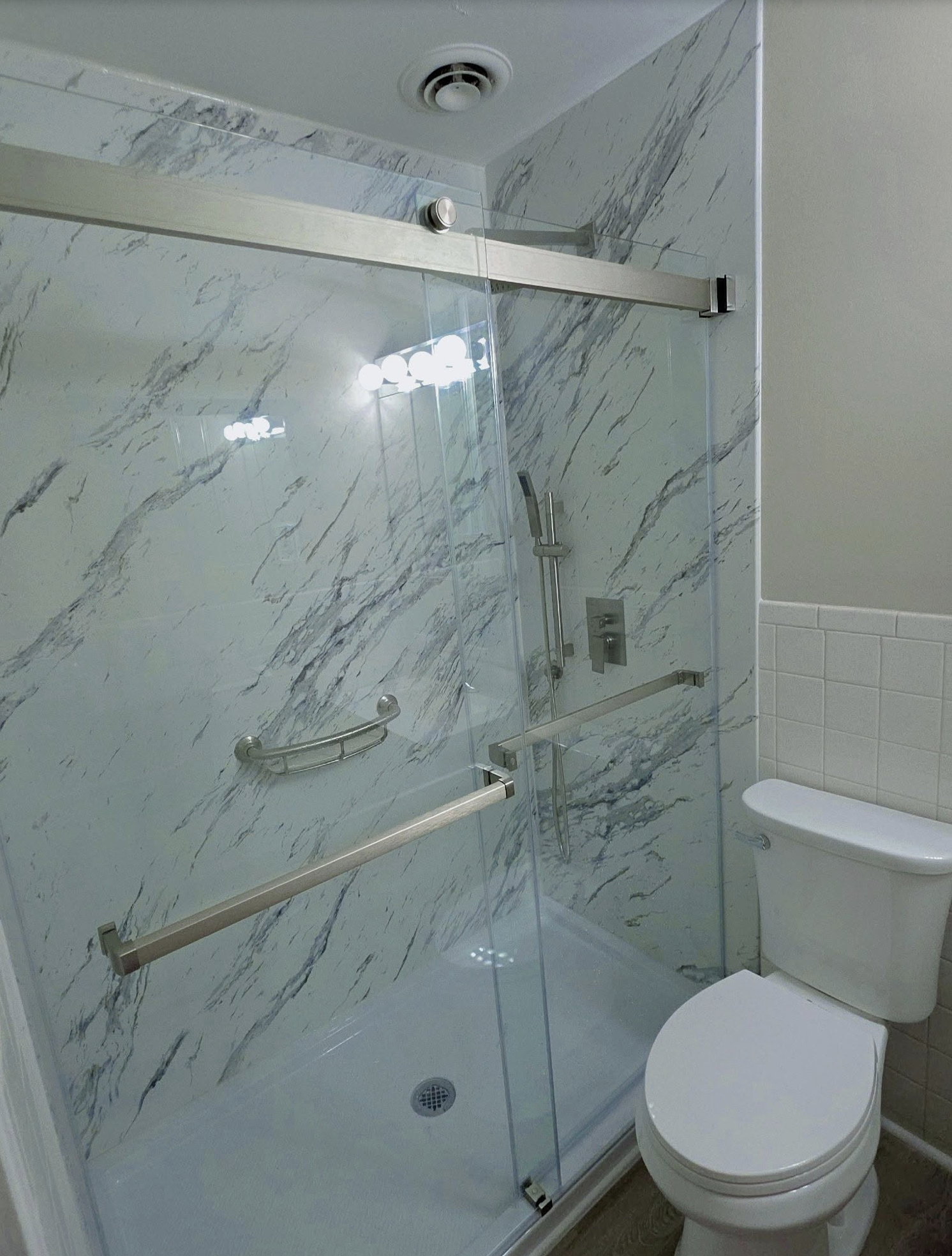 A bathroom with marble walls, a glass shower enclosure, a showerhead, grab bars, and a white toilet.