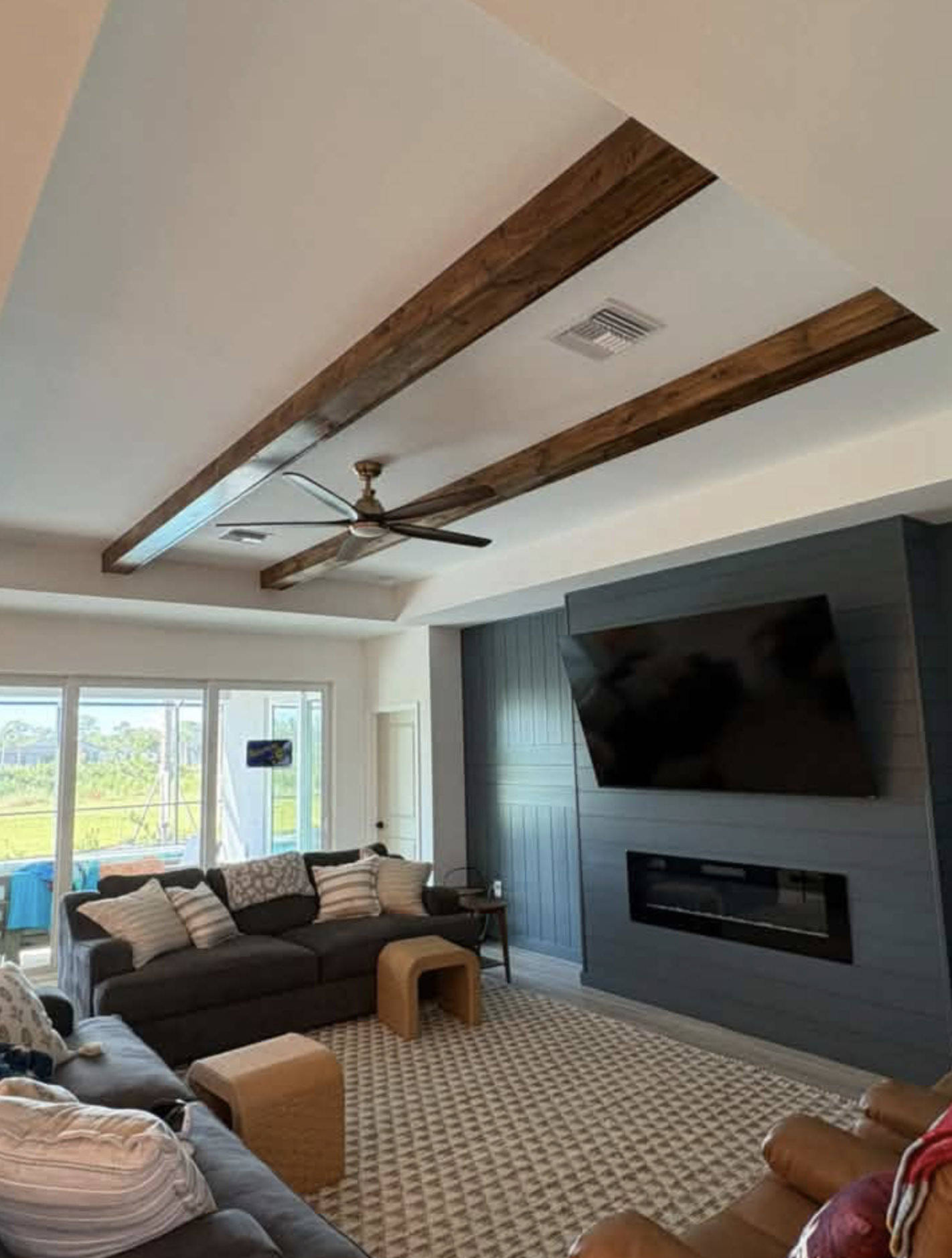 Living room with a dark sofa and an armchair, a patterned area rug, and a blue wall with a mounted TV and a fireplace. Large windows in the background reveals a porch and green outdoor landscape. The ceiling has wooden beams and a ceiling fan.