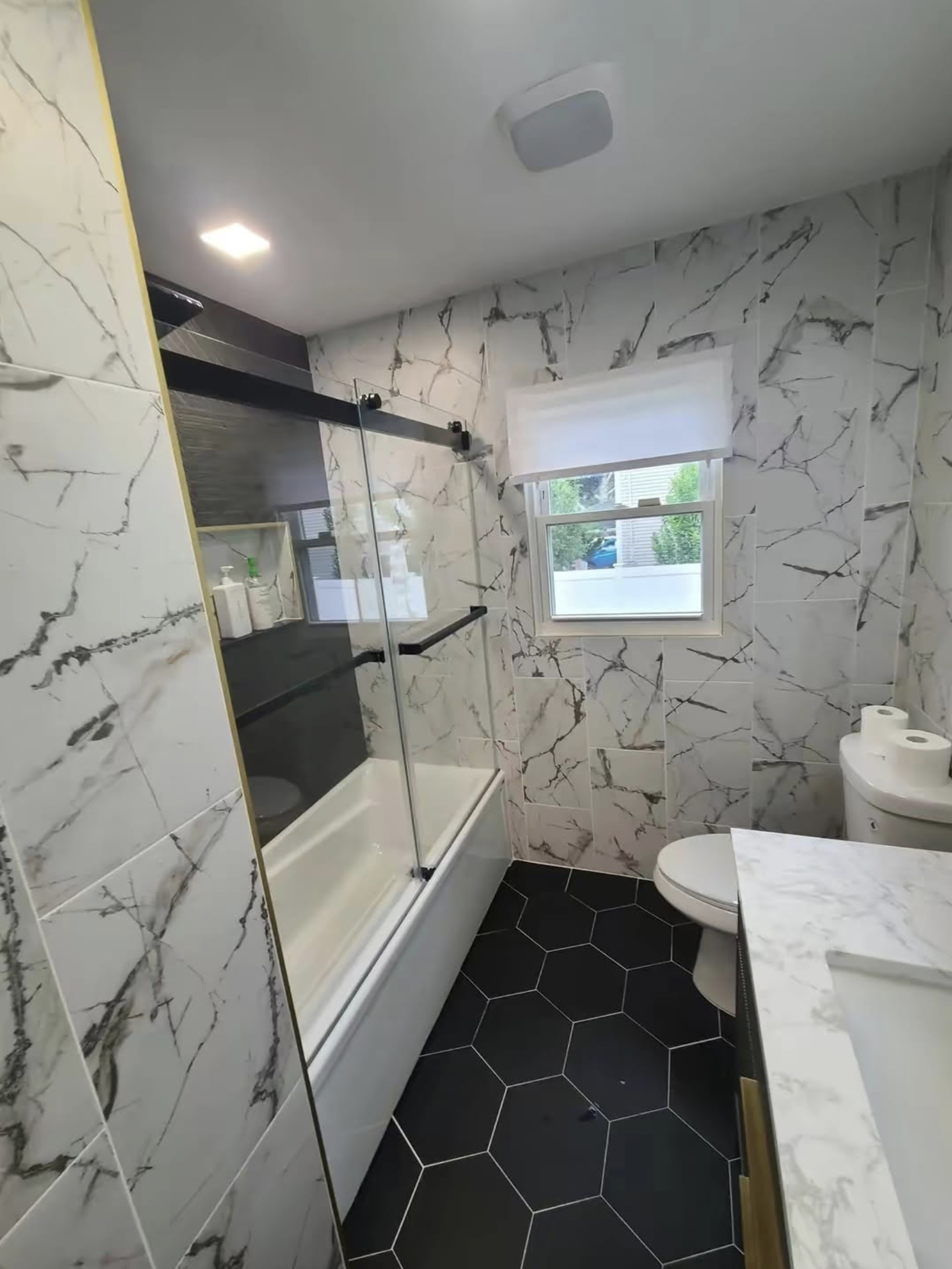 A modern bathroom with marble wall tiles, a glass shower enclosure, a window with a blind, and a toilet with a roll of toilet paper on the floor.