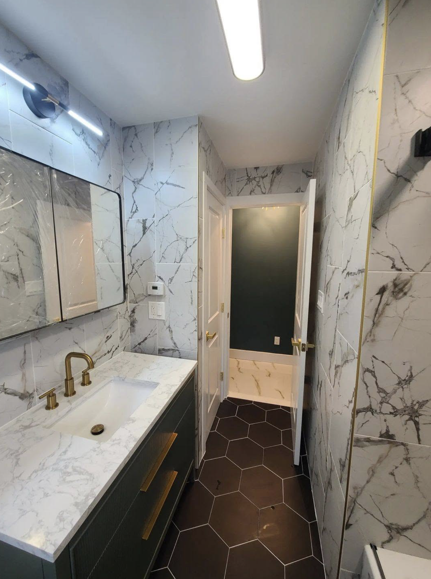 A modern bathroom with white marble walls and black hexagonal floor tiles. It features a black vanity with gold handles, a white marble countertop, a gold faucet, and a large mirror. There is an open door leading to another room, and the lighting inc
