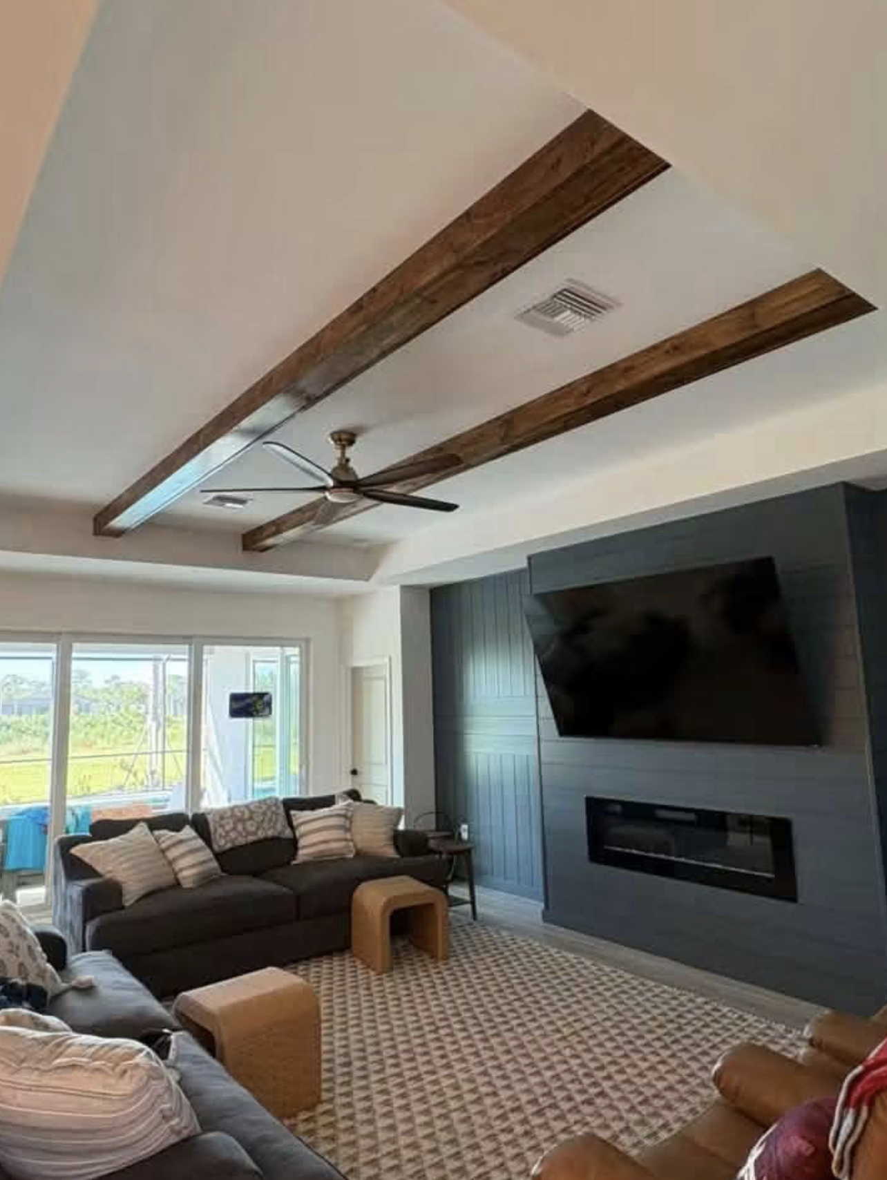 Living room with a ceiling fan, wooden beams, a large wall-mounted television, a dark sofa with pillows, a smaller armchair, a patterned rug, and sliding glass doors letting in natural light.