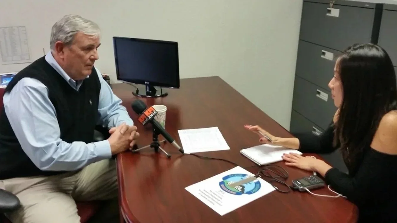 A man and woman having an interview at a desk, with a microphone, papers, and a computer monitor.