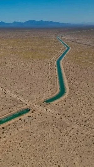 Aerial view of a winding canal through a desert landscape with mountains in the background.