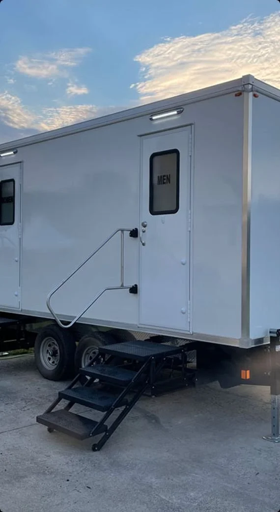 A white mobile restroom trailer with steps leading up to the door labeled 'MEN,' parked on a concrete surface under a partly cloudy sky.
