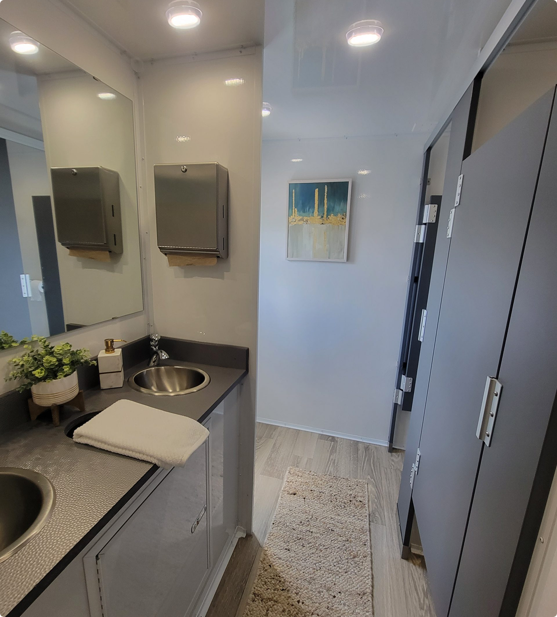 A small laundry room with a gray cabinet washer and dryer, a narrow beige rug, a piece of abstract wall art, and a small sink with a plant and soap dispenser.