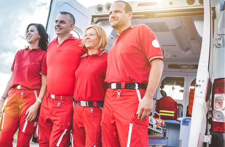 Four emergency medical responders stand outside an ambulance, smiling and looking into the distance.