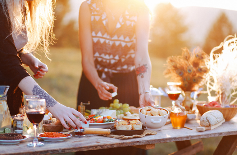 People gathered around a rustic outdoor table sharing a meal at sunset, with food, drinks, and decorative peacock feathers.