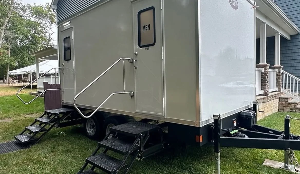 Portable restroom trailer with two separate entrances labeled 'MEN' and 'WOMEN', set on a grassy area near a house with stairs and porch.