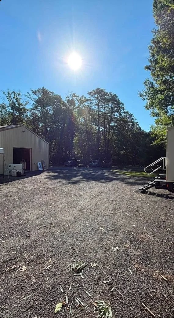 Sun shining brightly over a gravel parking lot with trees in the background and two buildings on either side.