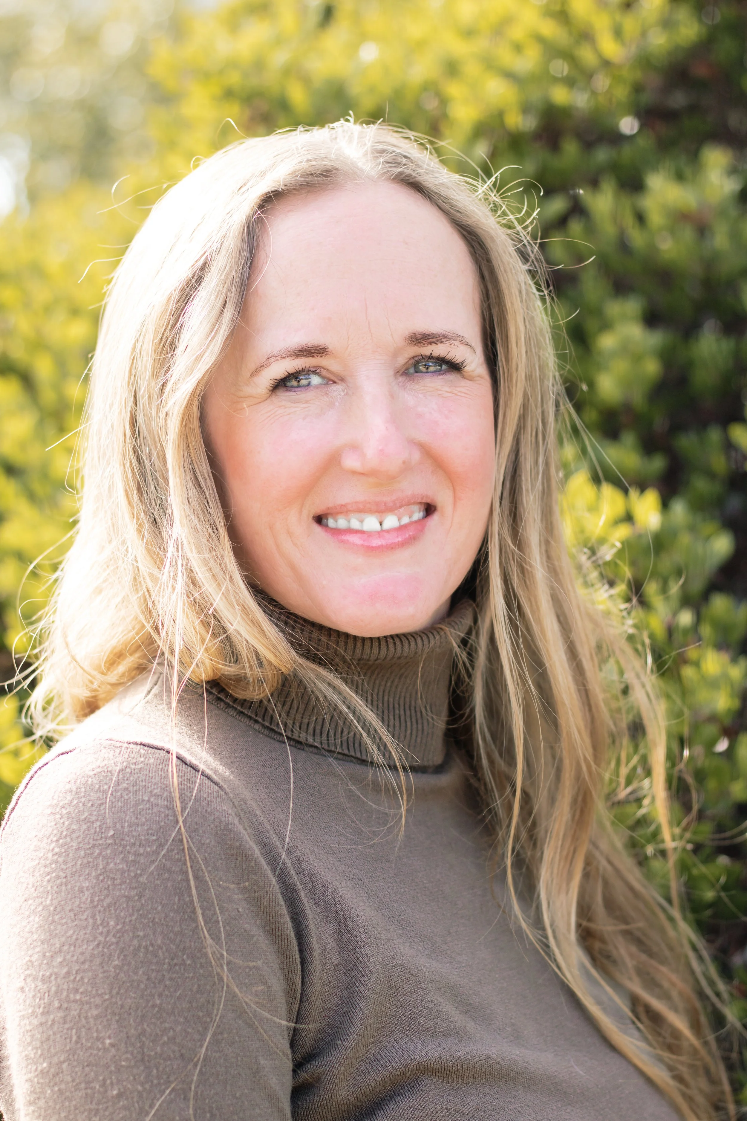 A smiling woman with long blonde hair outdoors with green foliage in the background.