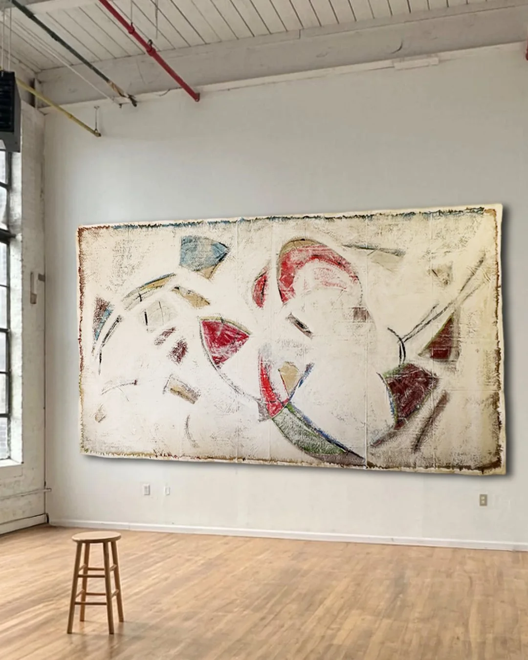 An abstract painting with colorful geometric shapes on a large canvas hung on a white wall in a gallery. A small wooden stool is on the wooden floor in front of the painting.