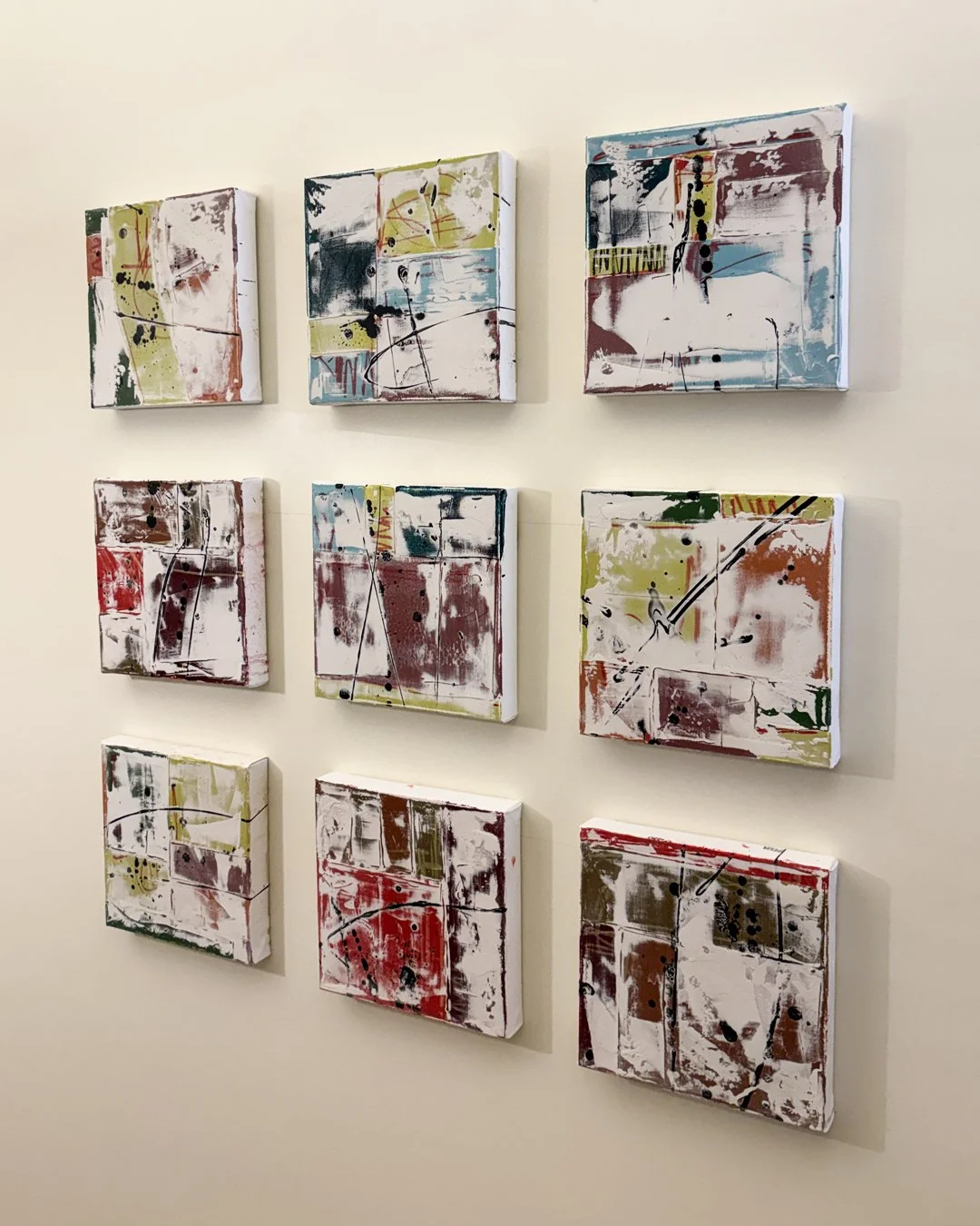 Nine abstract mixed media paintings arranged in a grid on a white wall, featuring layered colors, splatters, and lines.