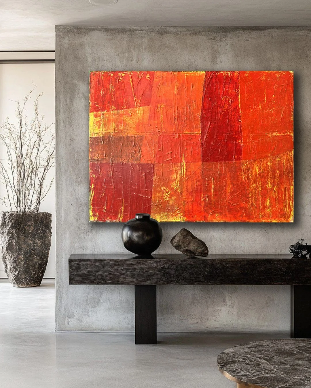 Modern interior with an abstract orange and red painting on a concrete wall, black vases and stones on a dark wooden console, a large potted plant in a textured pot in the background.