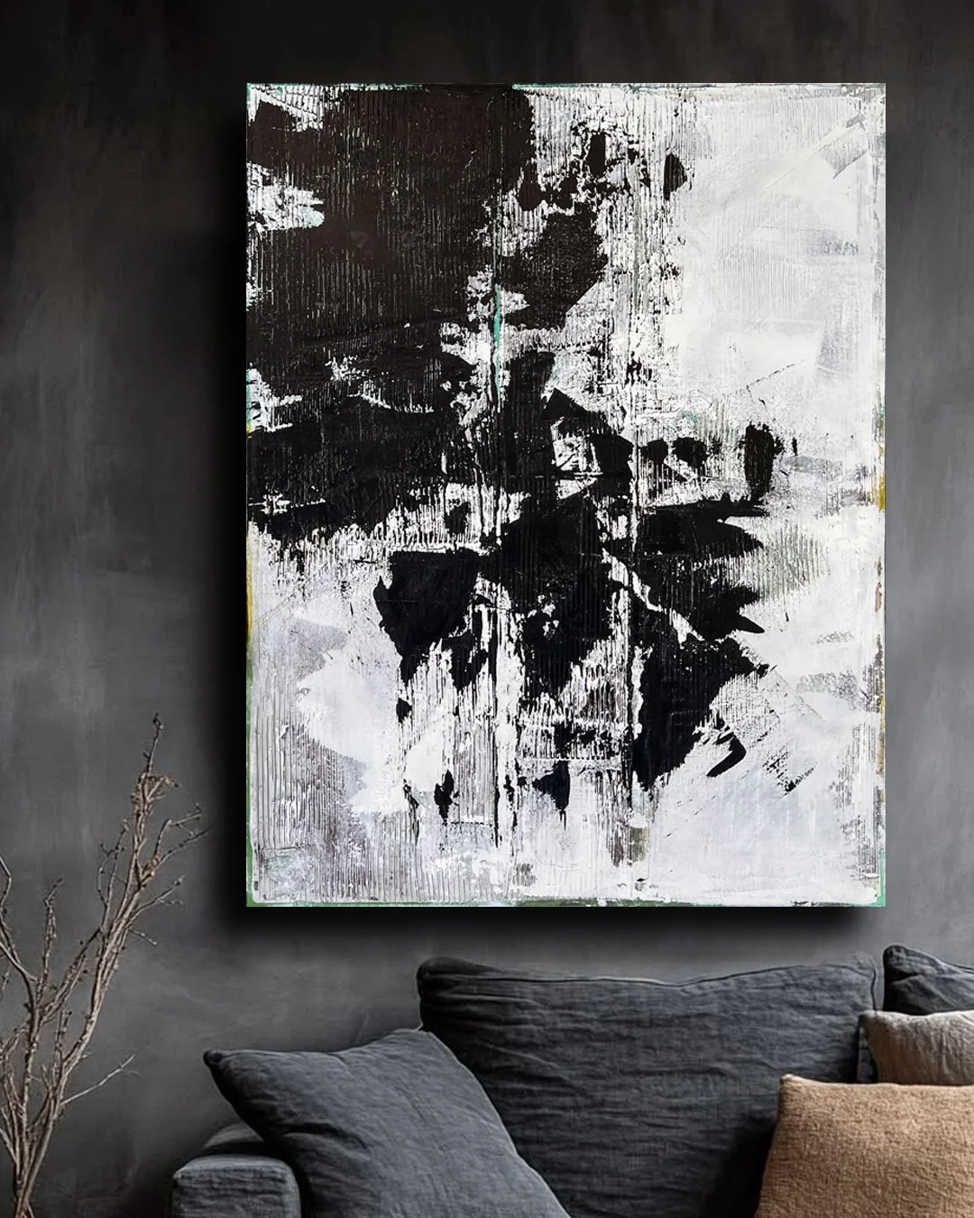 Abstract black and white textured painting hanging on a dark gray wall above a gray sofa and beige cushions, with a dried plant in a vase nearby.