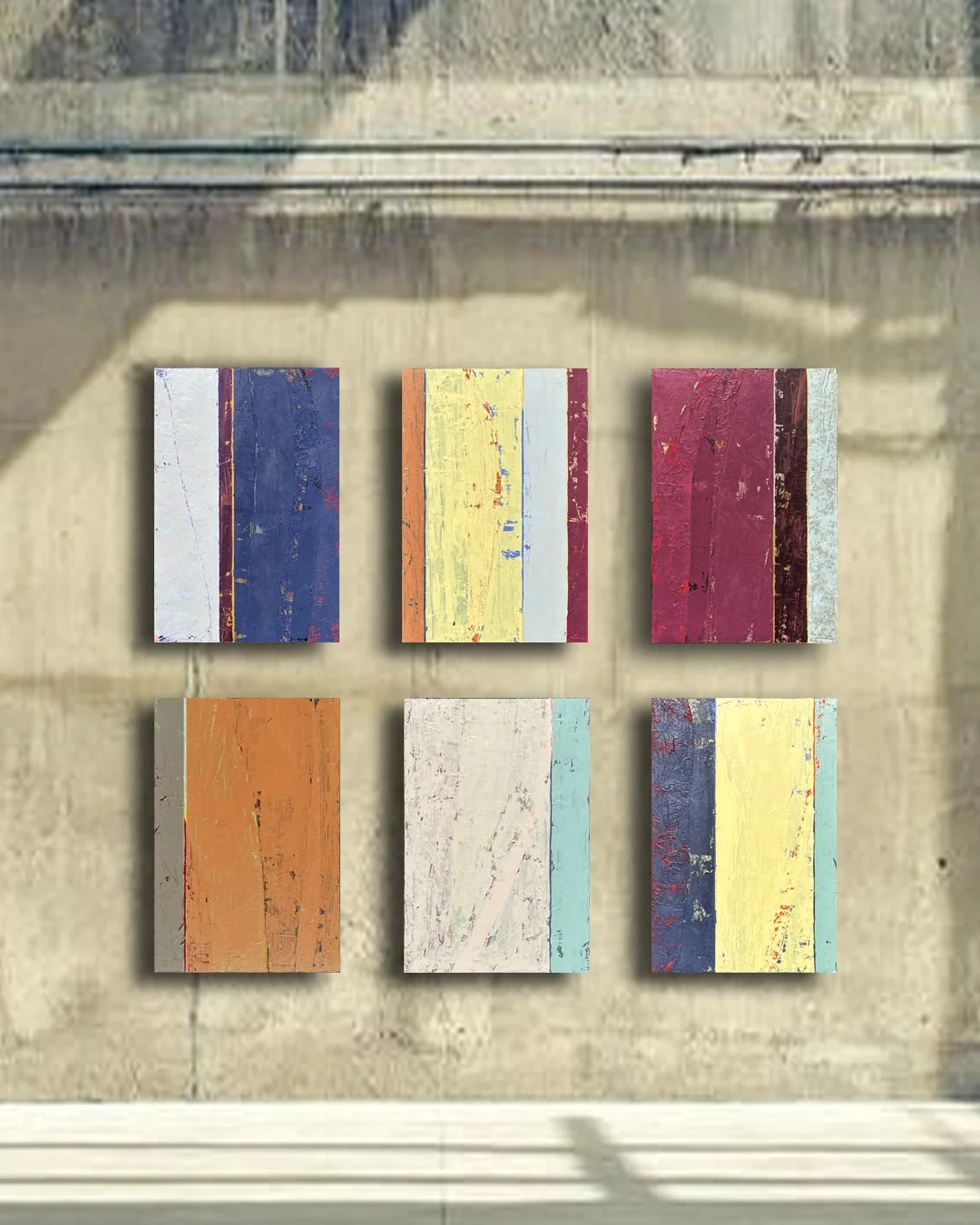 Six abstract paintings with colorful, distressed textures arranged in two rows on a brick wall.