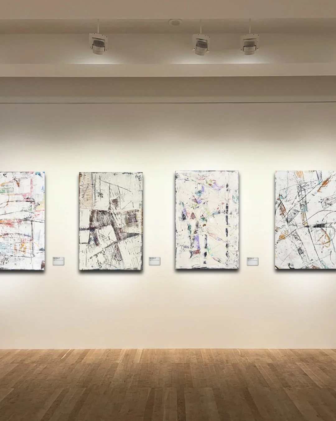 Art gallery wall with four abstract paintings, each with a mix of lines and colors on a white background, illuminated by ceiling spotlights.